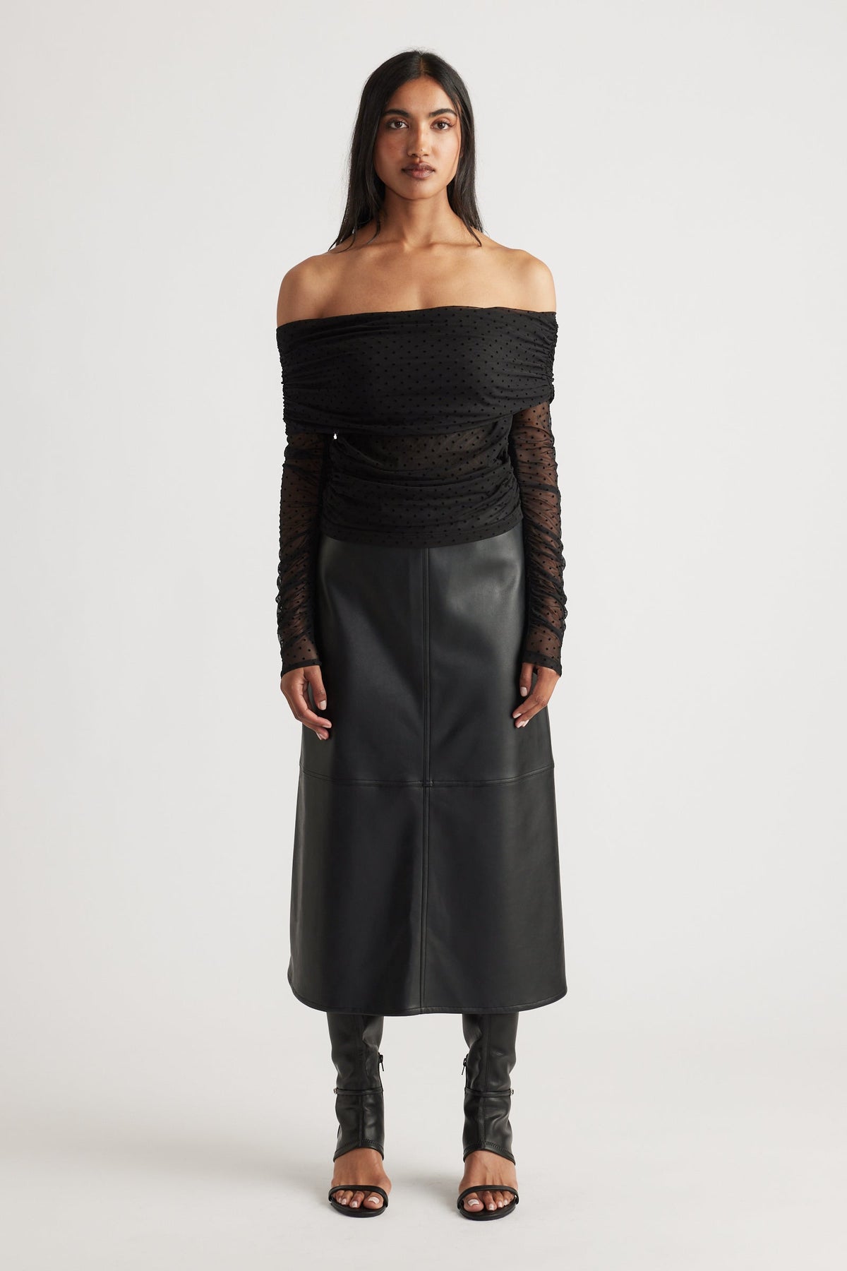 Woman wearing a black off-shoulder ruched mesh top paired with a high-waisted faux leather midi skirt and open-toe heels