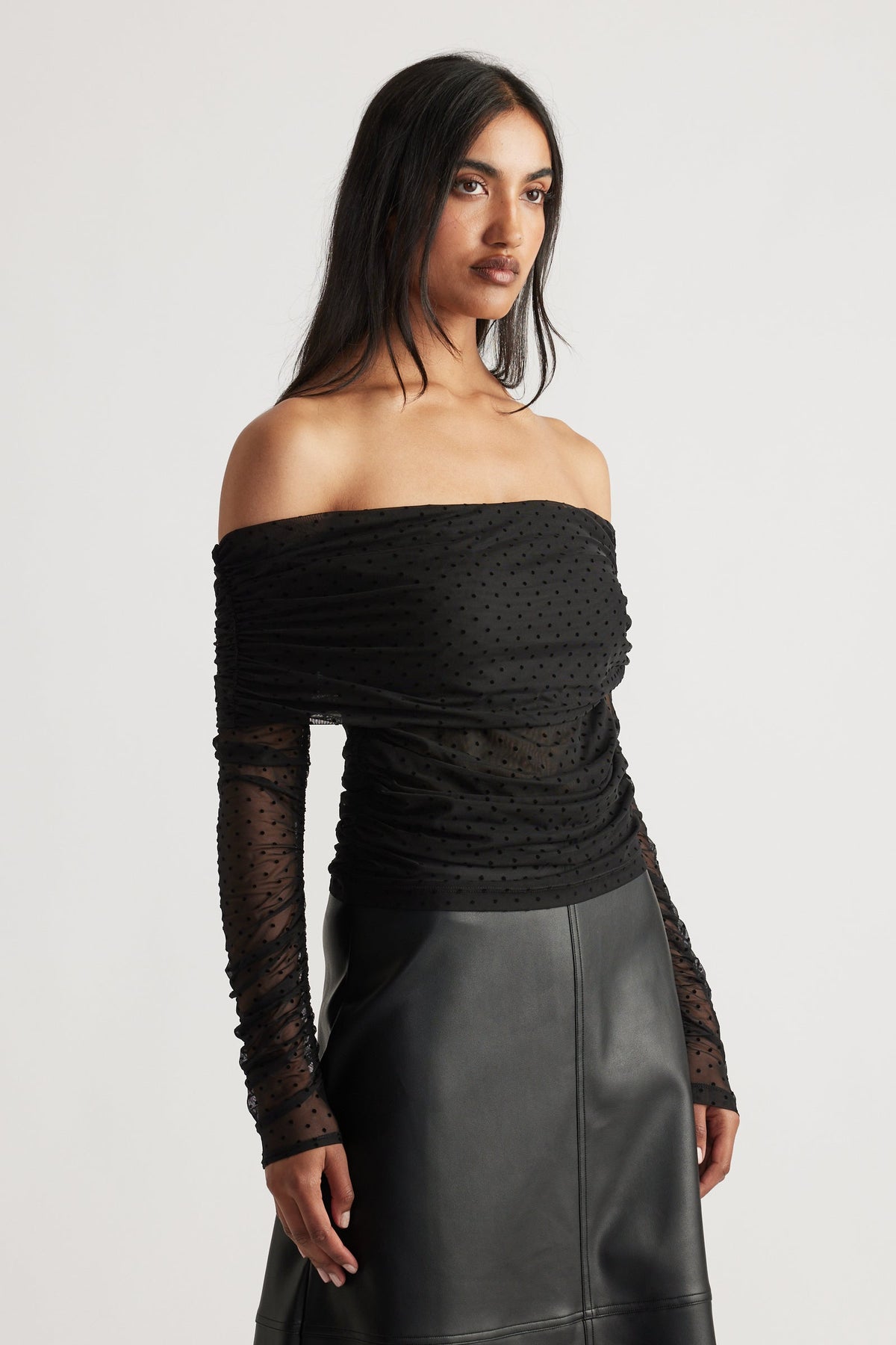 Woman wearing a fitted off-shoulder black mesh top with ruched detailing and long sheer sleeves paired with a leather skirt