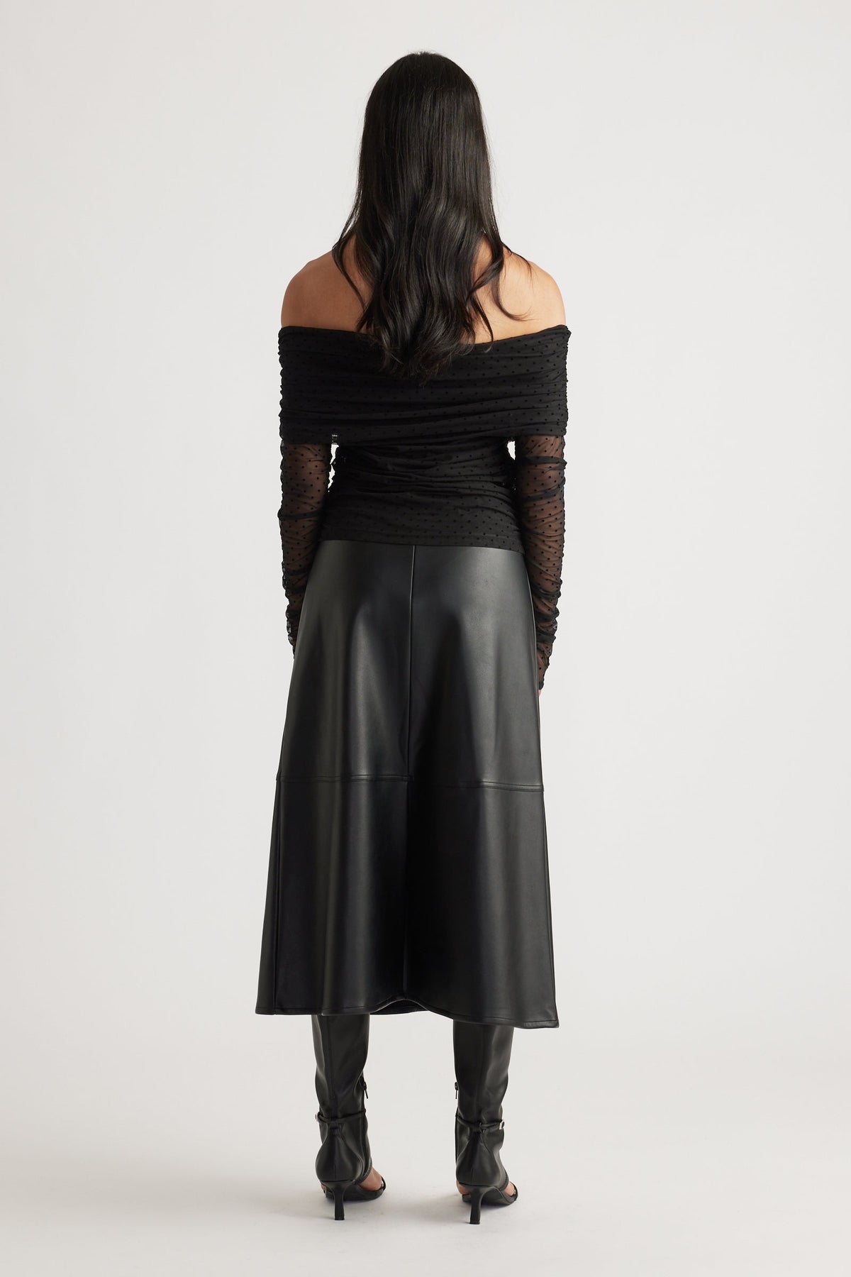 Back view of a woman wearing a fitted off-shoulder ruched mesh top with sheer sleeves, paired with a black midi skirt and heels