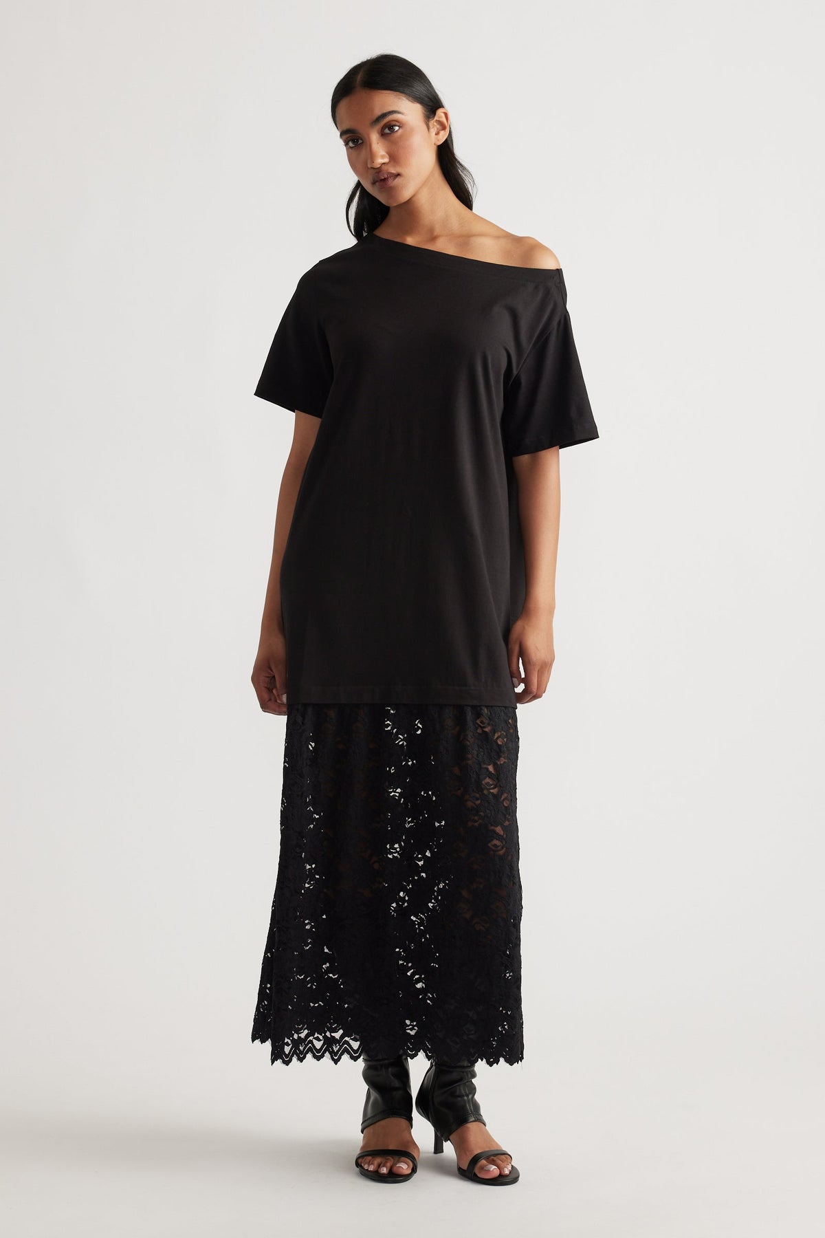 Woman wearing an off-shoulder black top layered over a long black lace skirt, paired with black open-toe heels.