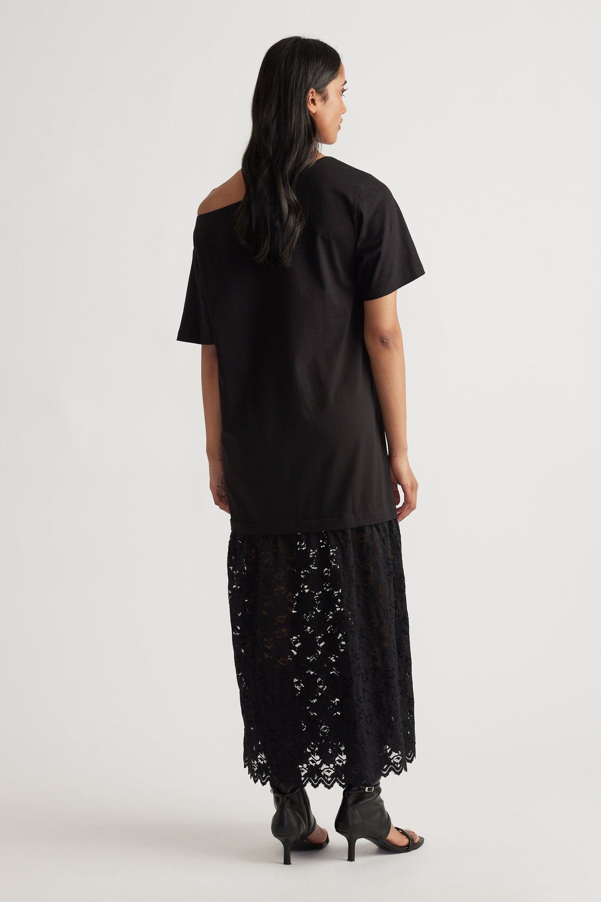 Back view of woman wearing a relaxed black top with an asymmetrical neckline over a sheer black lace maxi skirt and heeled sandals