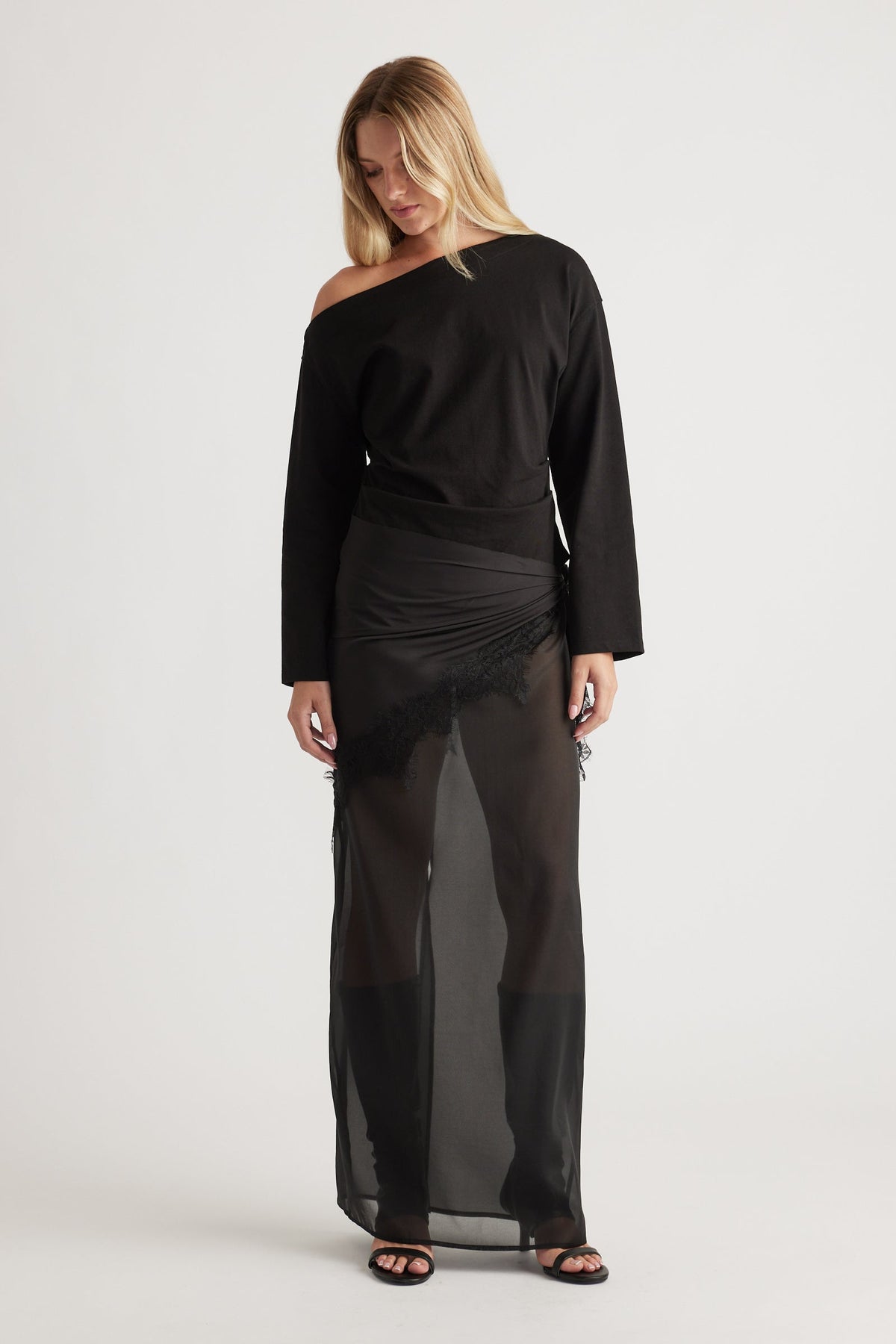 Woman wearing an off-shoulder black long sleeve top paired with a sheer black maxi skirt featuring lace trim details