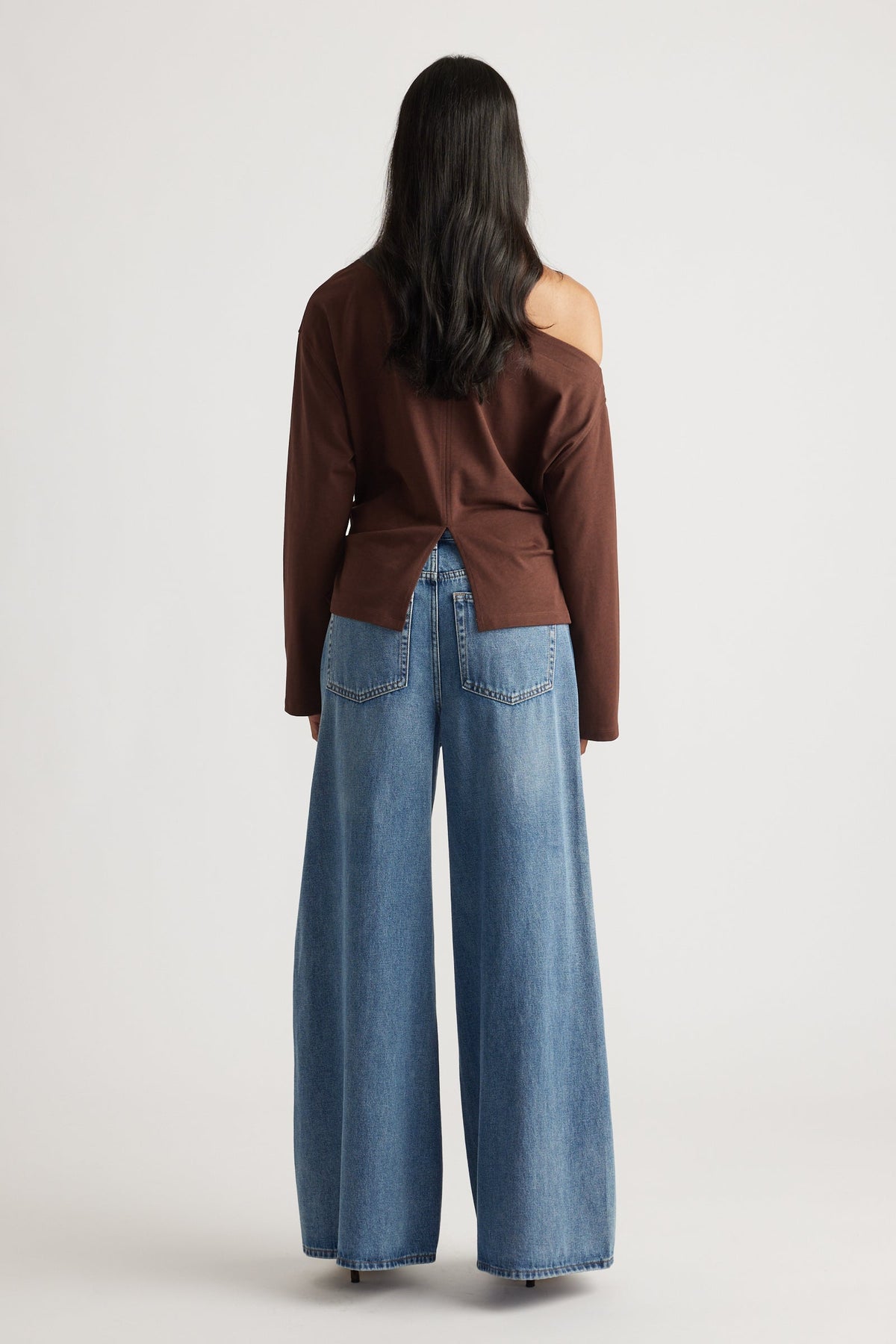 Back view of a woman wearing a brown off-shoulder top with a back slit, paired with wide-leg blue denim jeans