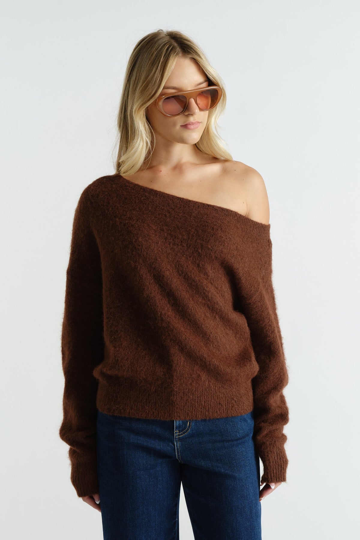 Woman wearing a soft brown off-shoulder knit sweater paired with dark blue jeans and brown-tinted sunglasses