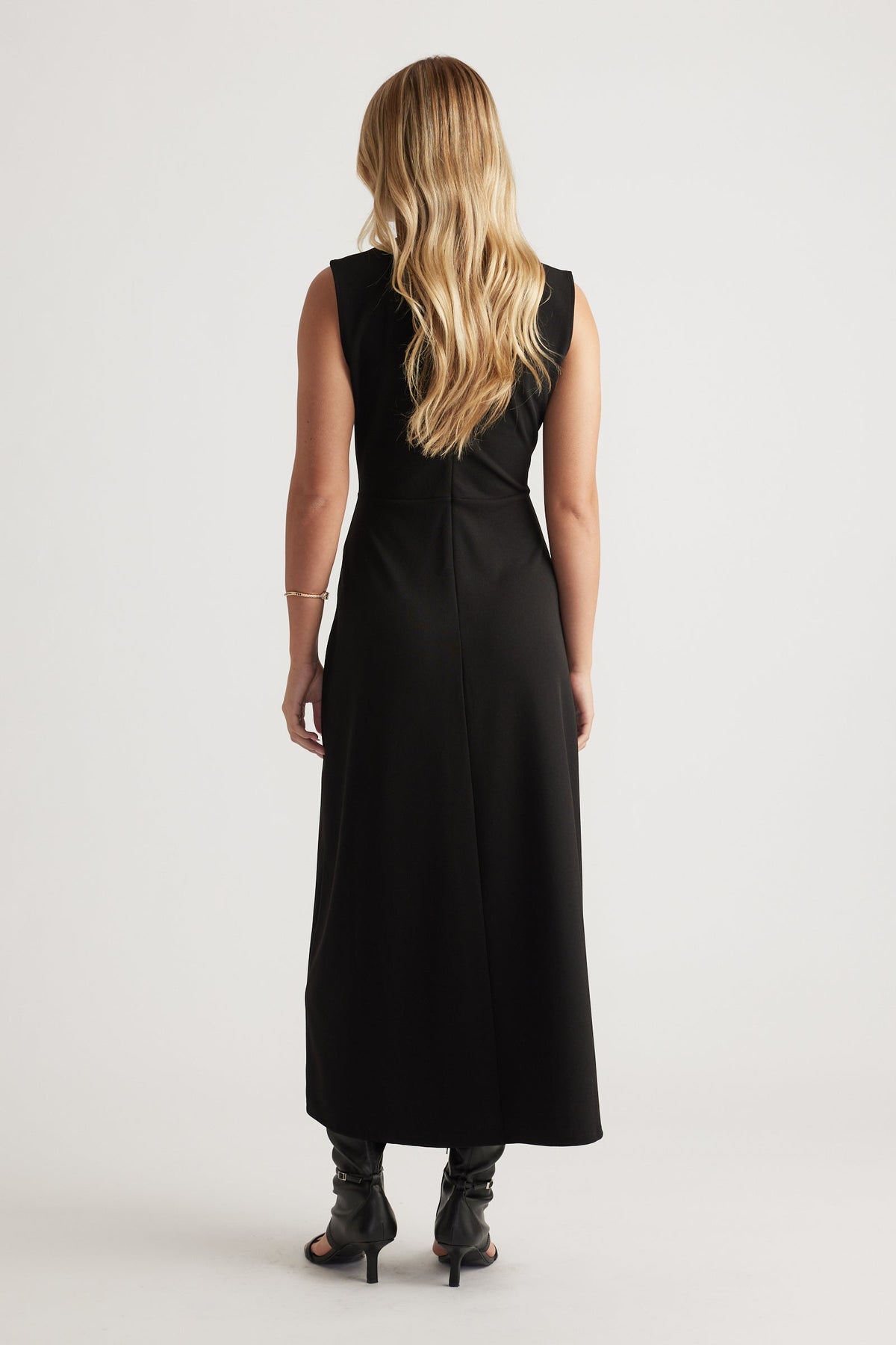 Back view of a woman wearing a sleeveless black midi dress paired with black heeled boots and loose blonde hair