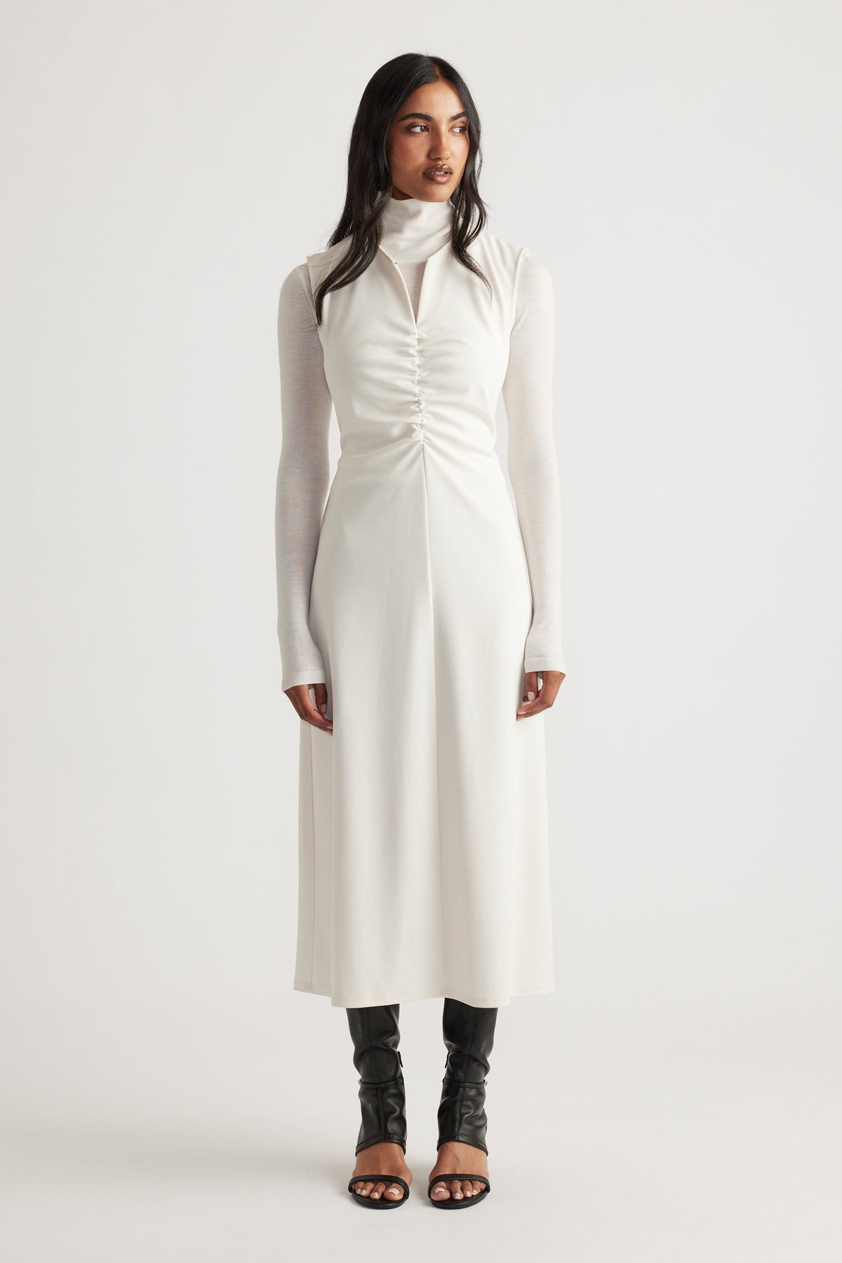 Woman wearing a white ruched midi dress layered over a fitted long-sleeve turtleneck and paired with black open-toe boots