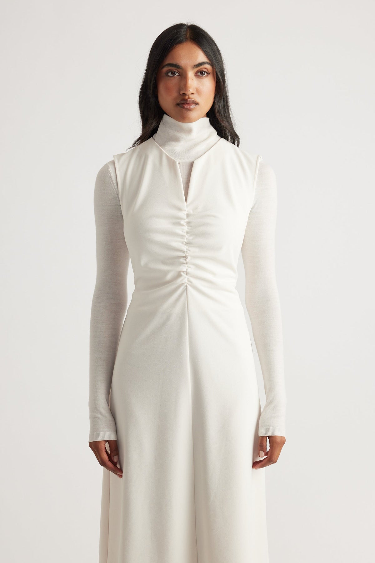 Woman wearing a white midi dress with ruched front detail layered over a fitted long sleeve turtleneck top
