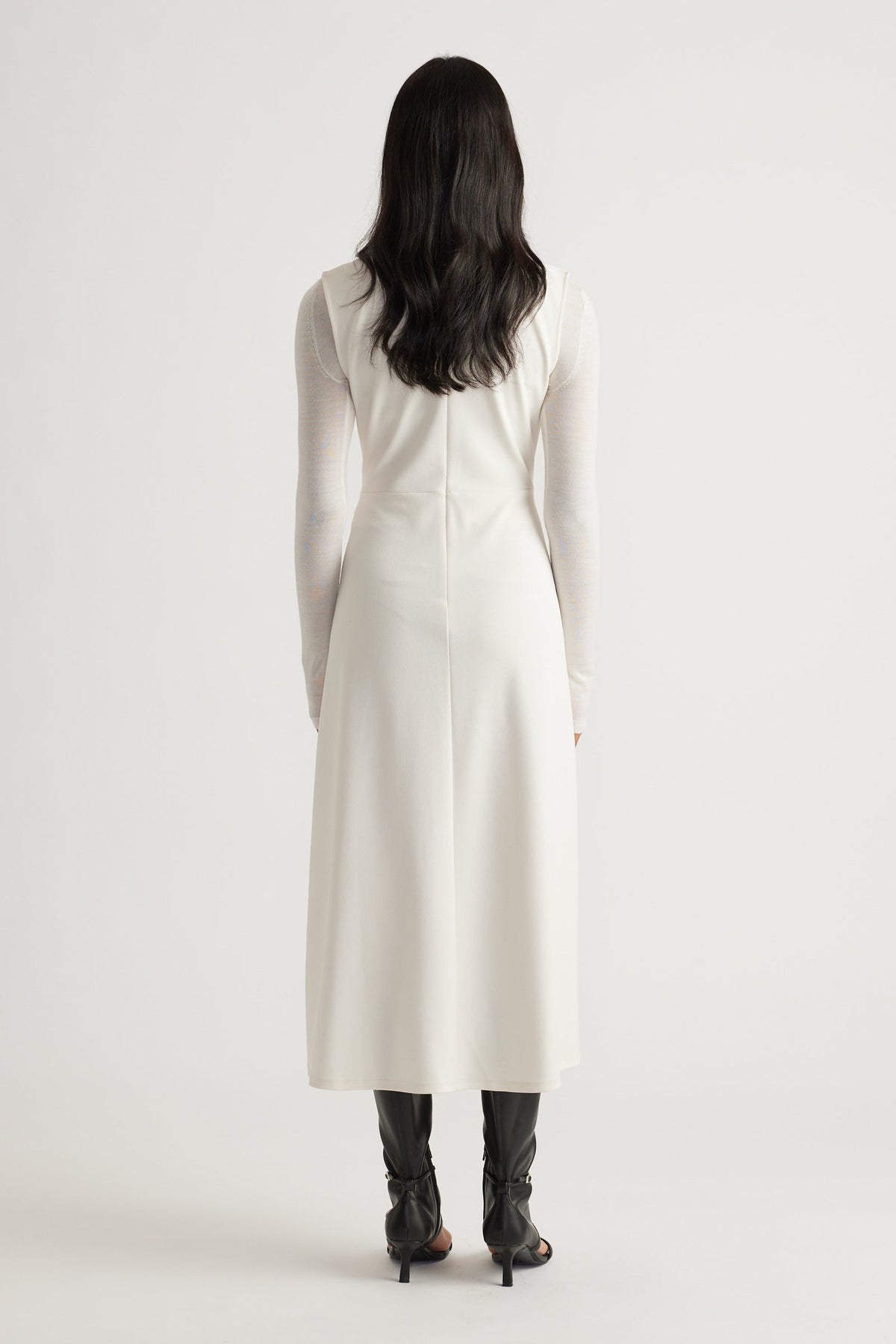 Back view of a sleeveless white midi dress worn over a sheer long-sleeve top paired with black heeled boots