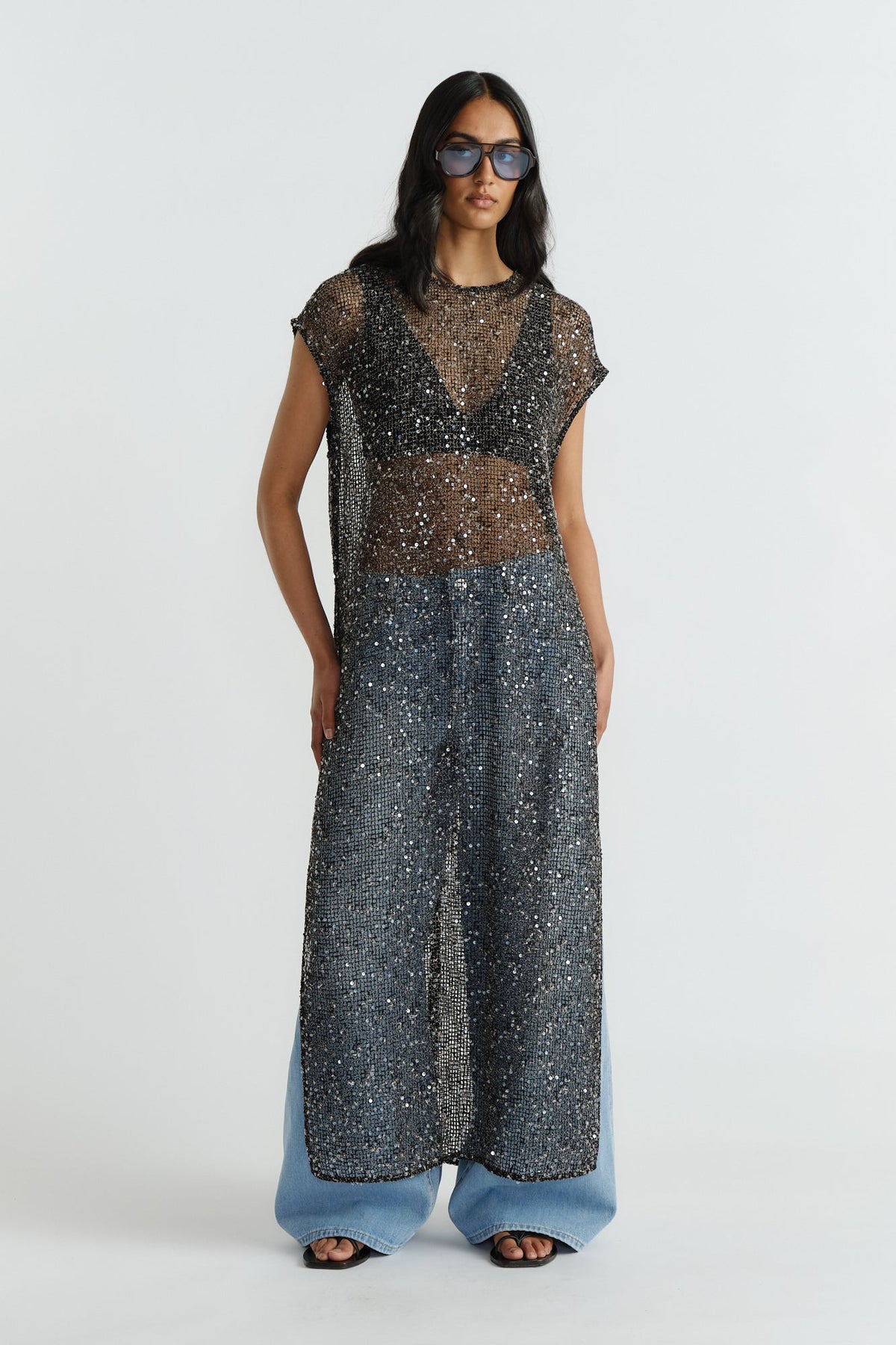 Model wearing a black sheer sequin dress layered over casual denim jeans and black sandals with oversized sunglasses.