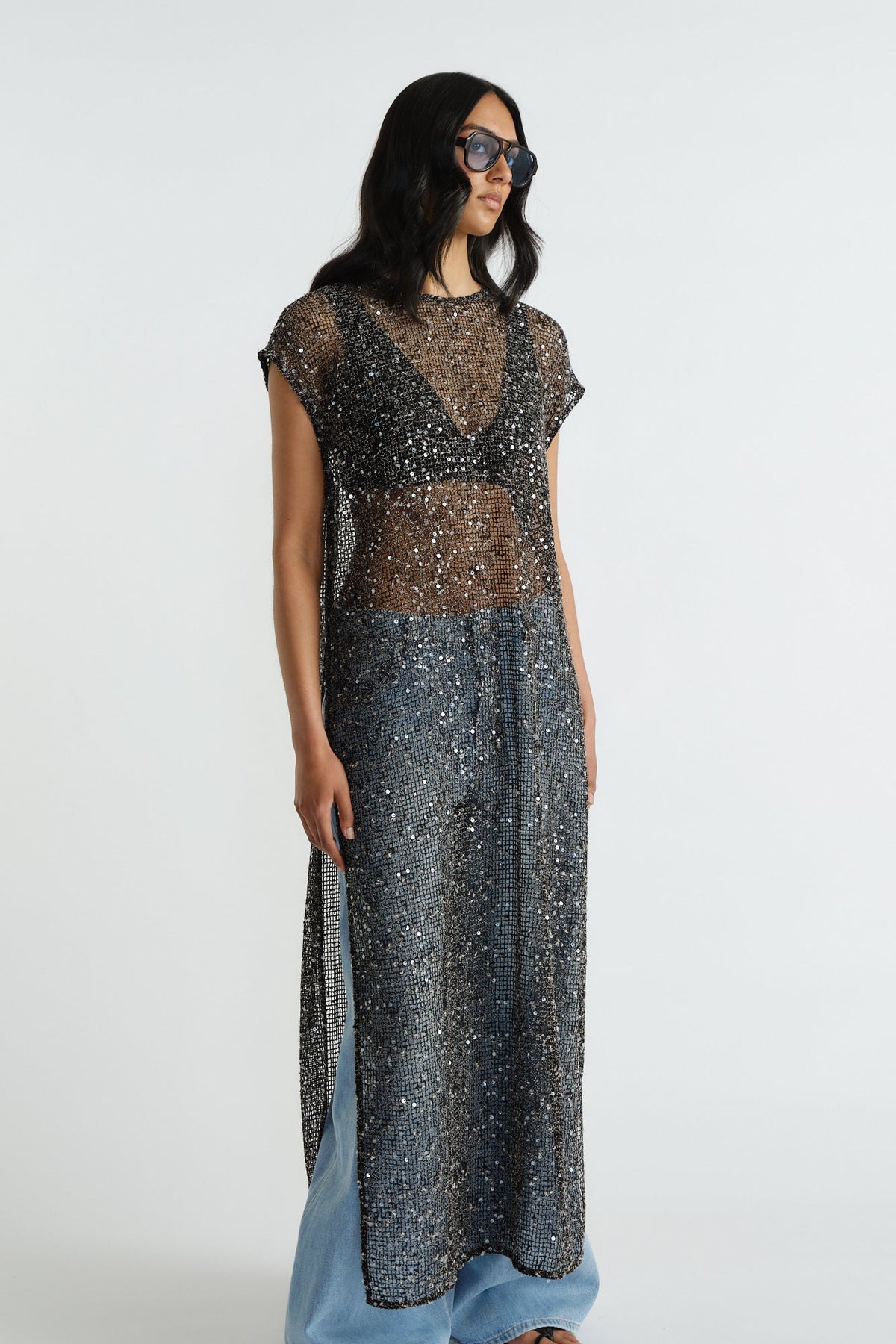 Woman wearing a long black sequin dress layered over casual denim jeans and a black bra top with sunglasses.