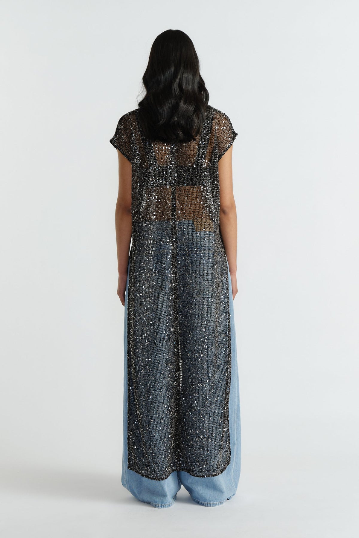 Back view of a woman wearing a long, sheer black sequin dress layered over wide-leg light blue jeans.