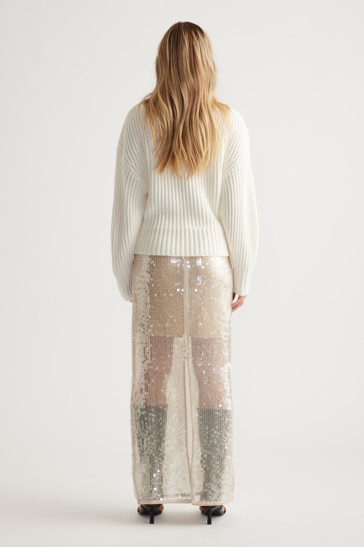 Woman wearing an off-white ribbed knit jumper paired with a shimmering sequin maxi skirt and black heels, shown from behind.