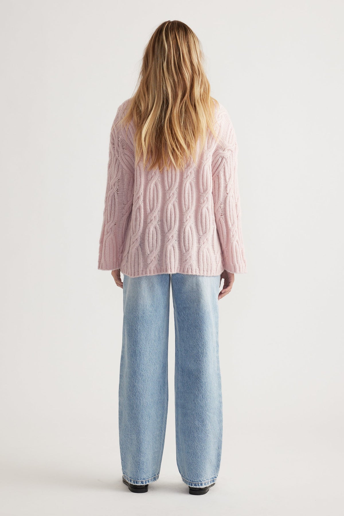 Woman with loose blonde hair wearing a soft pink cable knit jumper paired with light wash wide-leg jeans from behind