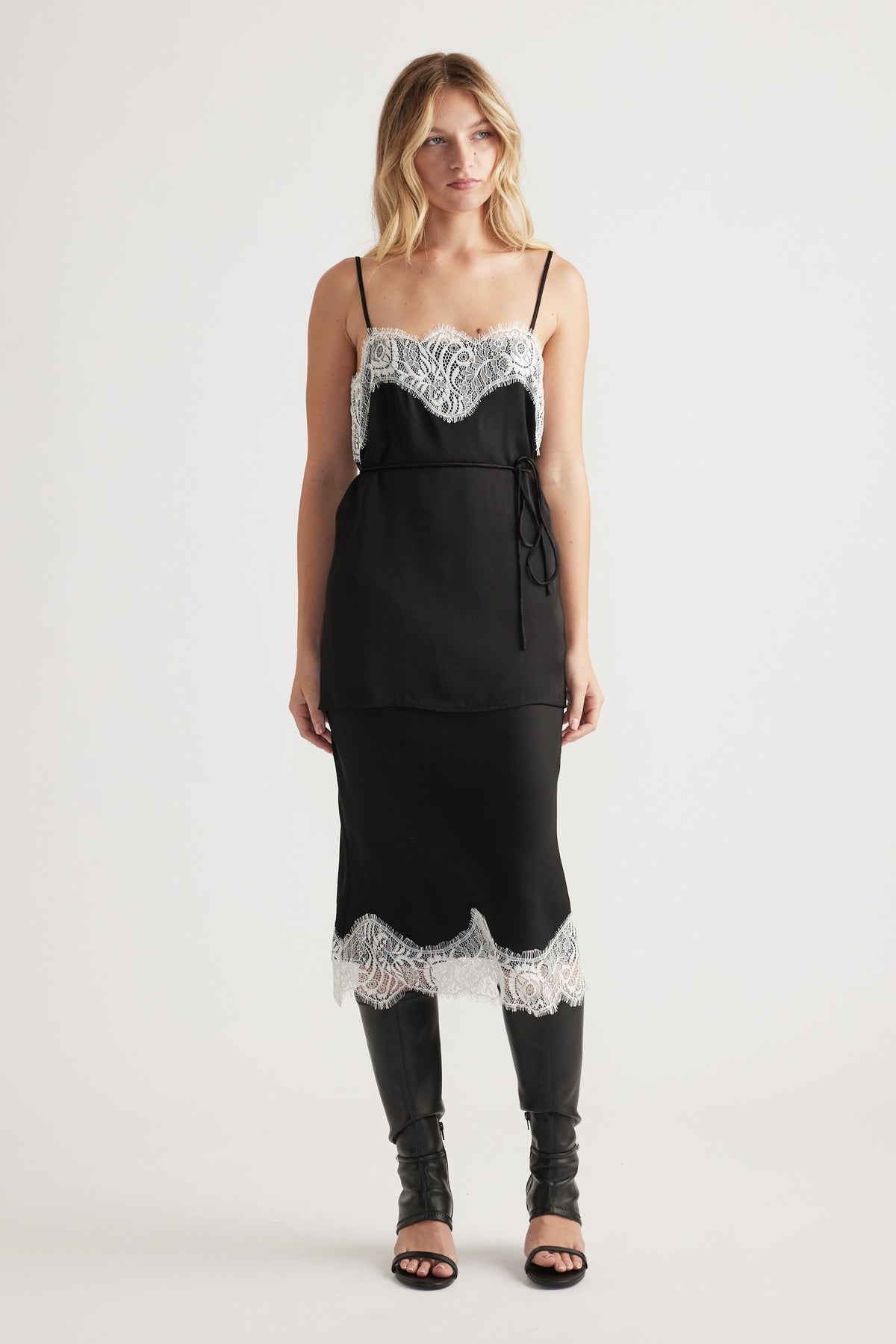 Woman wearing a black slip dress with white lace trim at neckline and hem, styled with tall black boots for an elegant look.