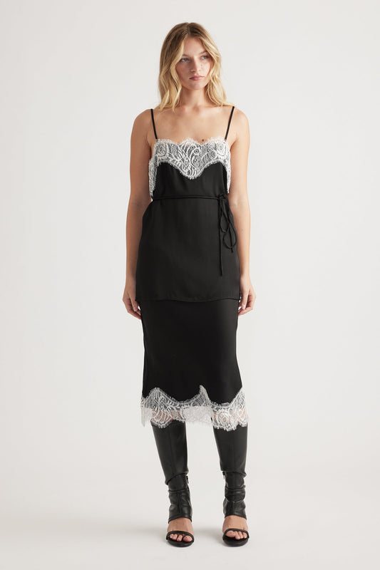 Woman wearing a black slip dress with white lace trim at neckline and hem, styled with tall black boots for an elegant look.