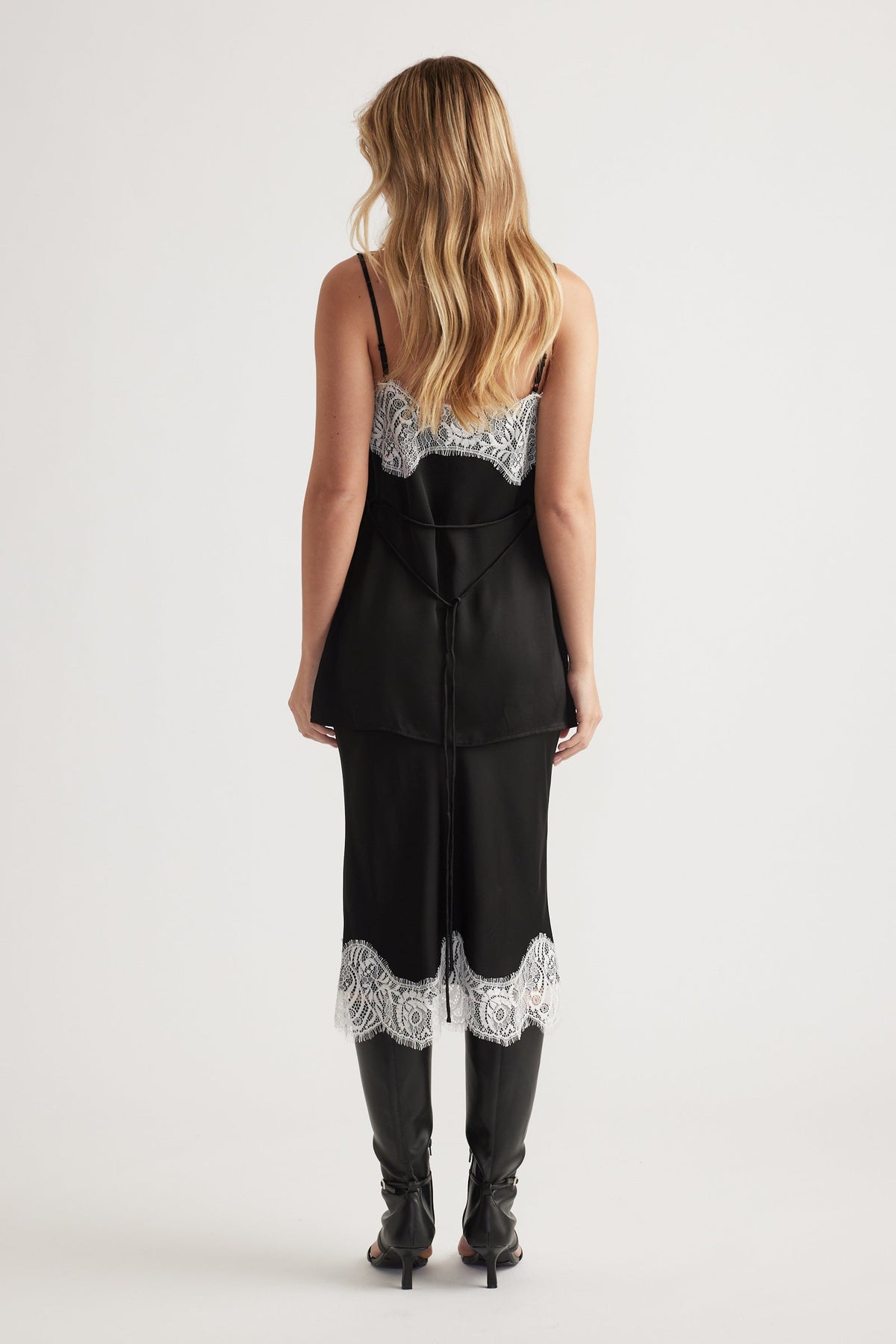 Woman wearing a black slip dress with white lace trim on the back and hem, paired with black high boots.