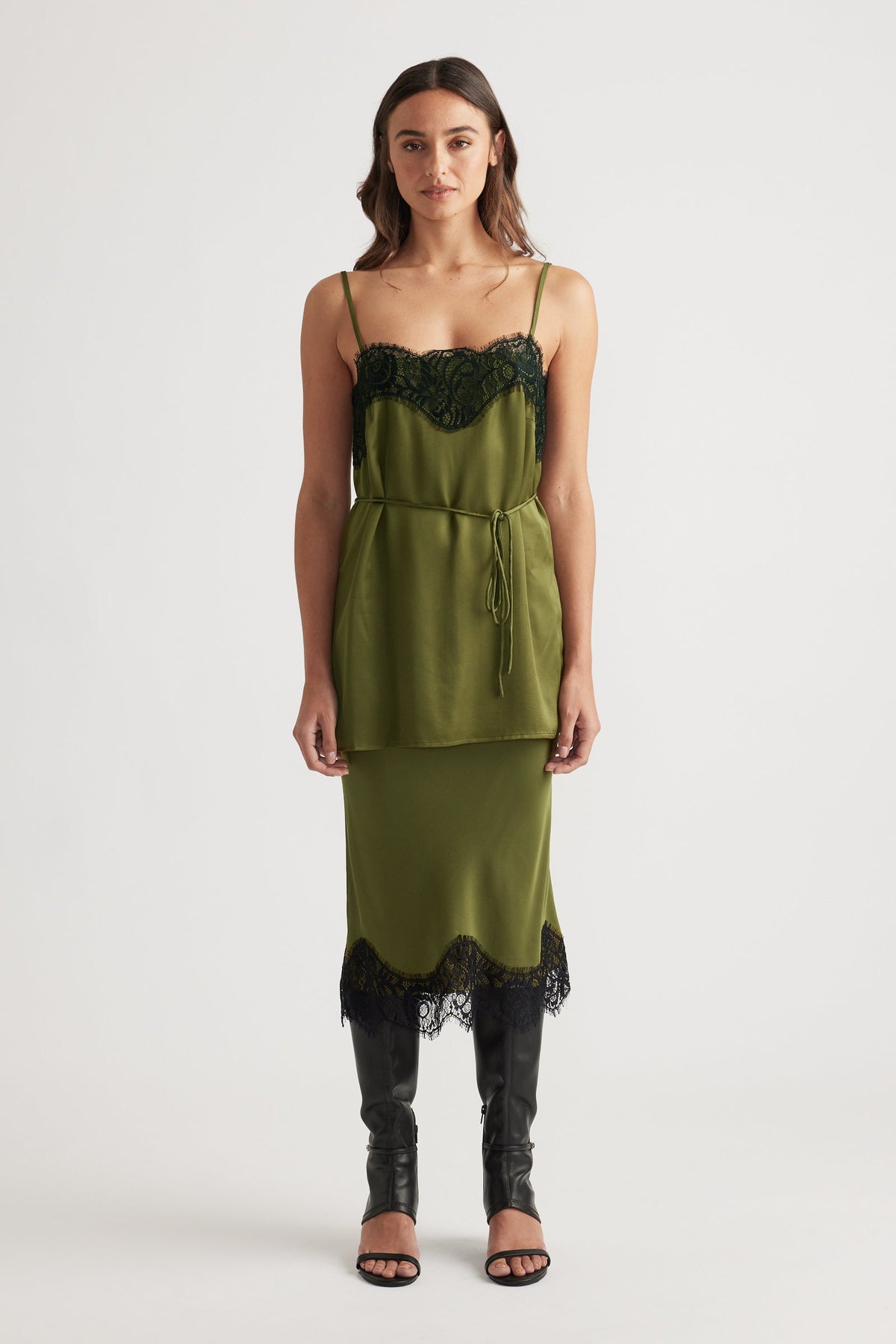 Front view of an olive green lace-trimmed slip dress layered with a matching tied waist top and black heeled boots