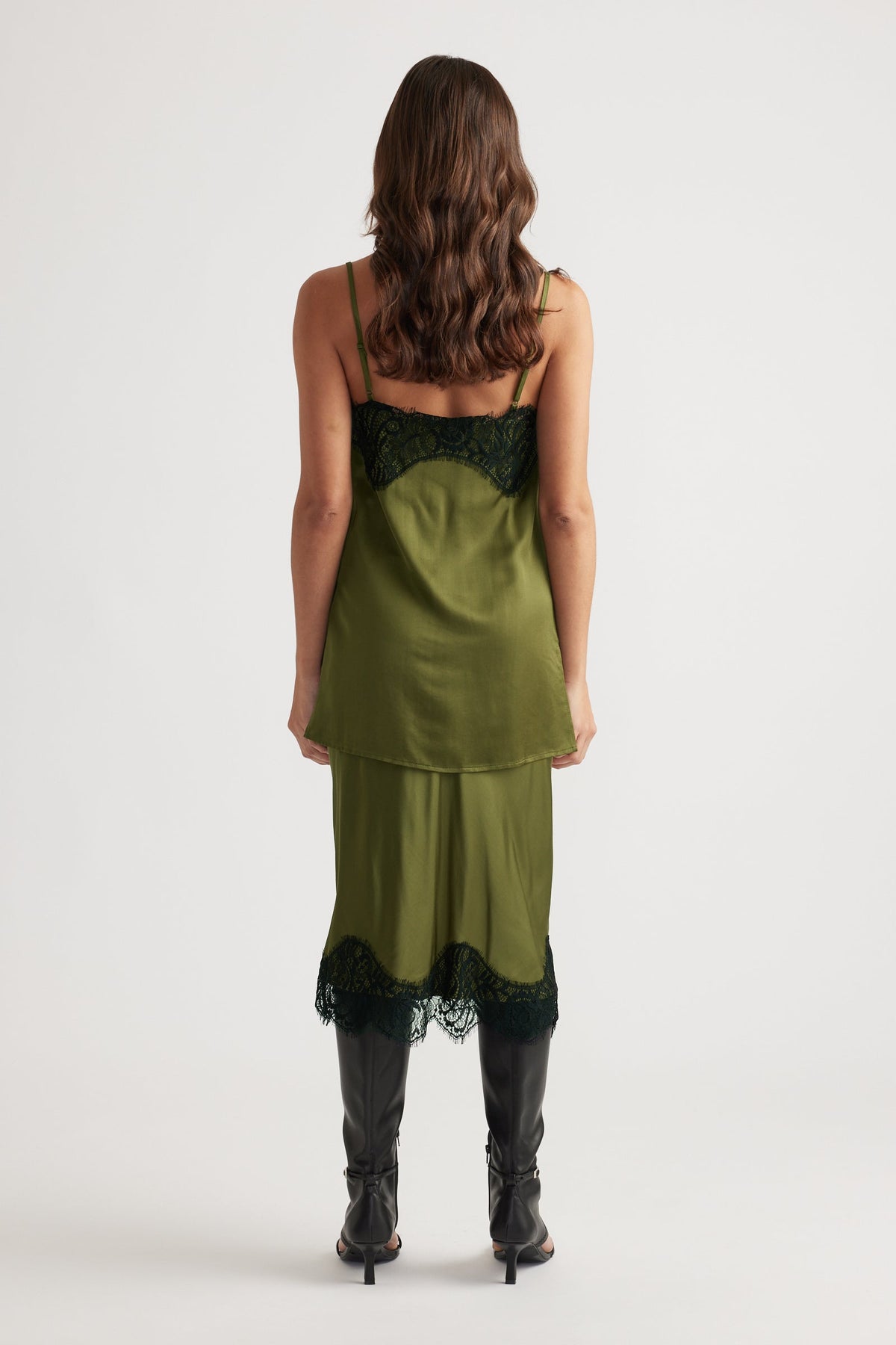 Back view of a silky olive green slip dress with black lace trim paired with knee-high black boots for a sleek look