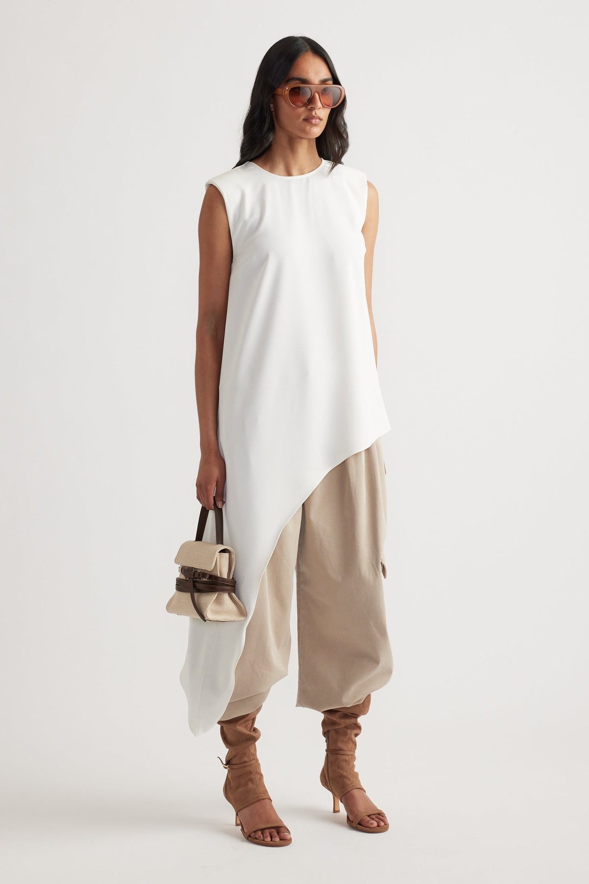 Woman wearing an ivory asymmetrical drape top paired with loose beige pants and tan heeled sandals carrying a small bag