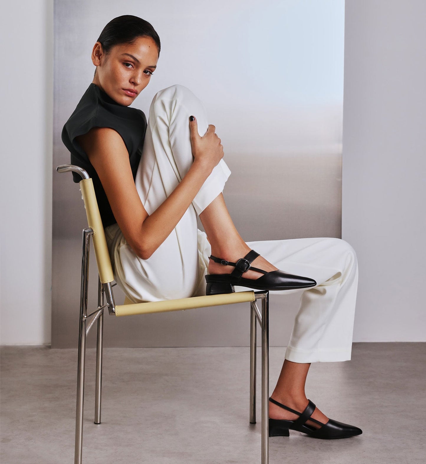 model sitting on a chair wearing black slingback flats and white pants with a black top