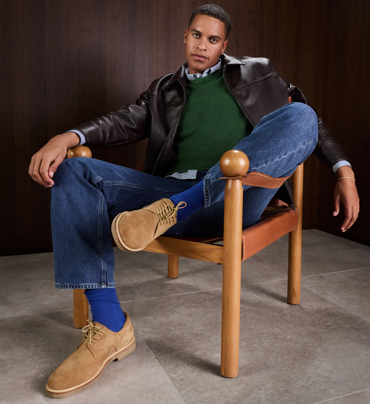Man sitting on a wooden chair wearing a leather jacket, green sweater, blue jeans, and tan suede shoes