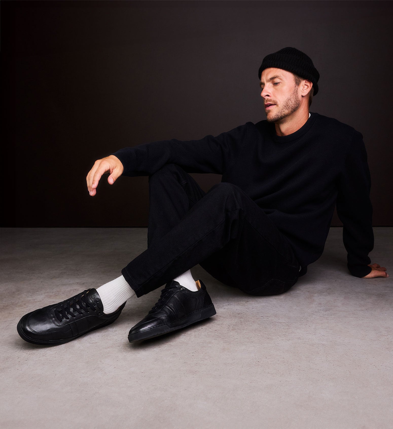 Man sitting on the floor wearing a black outfit with black shoes and a black beanie.