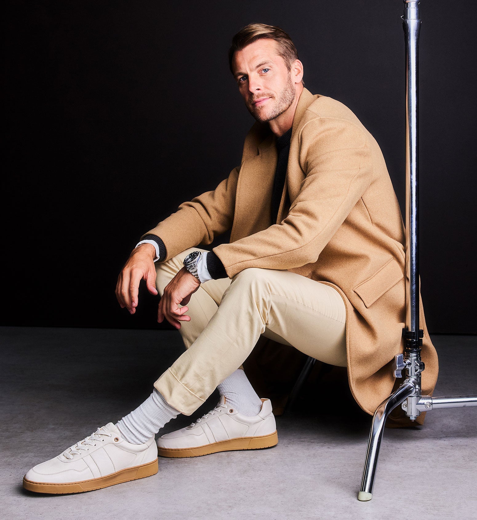 Man in beige coat and pants sitting on a gray floor wearing white sneakers with a black background