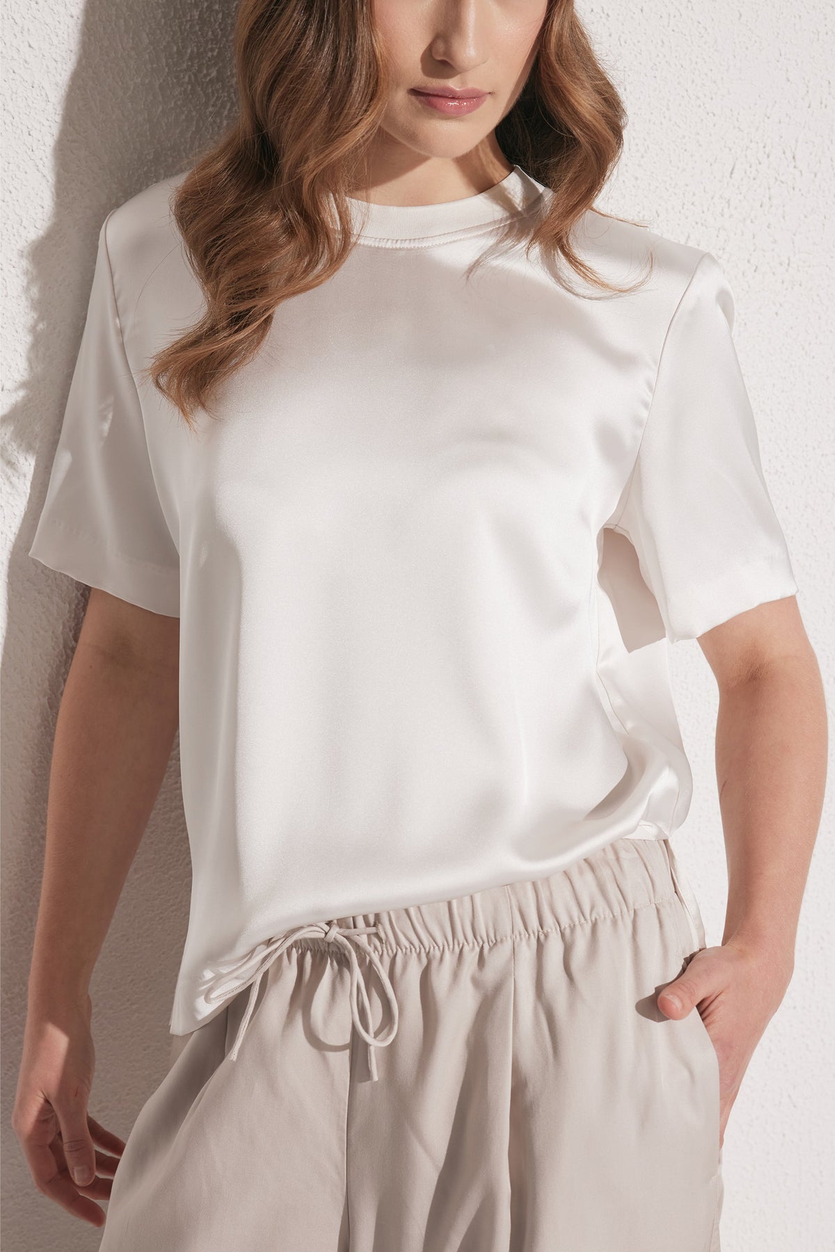 Woman wearing a smooth white satin tee with short sleeves paired with light beige drawstring pants against a textured wall