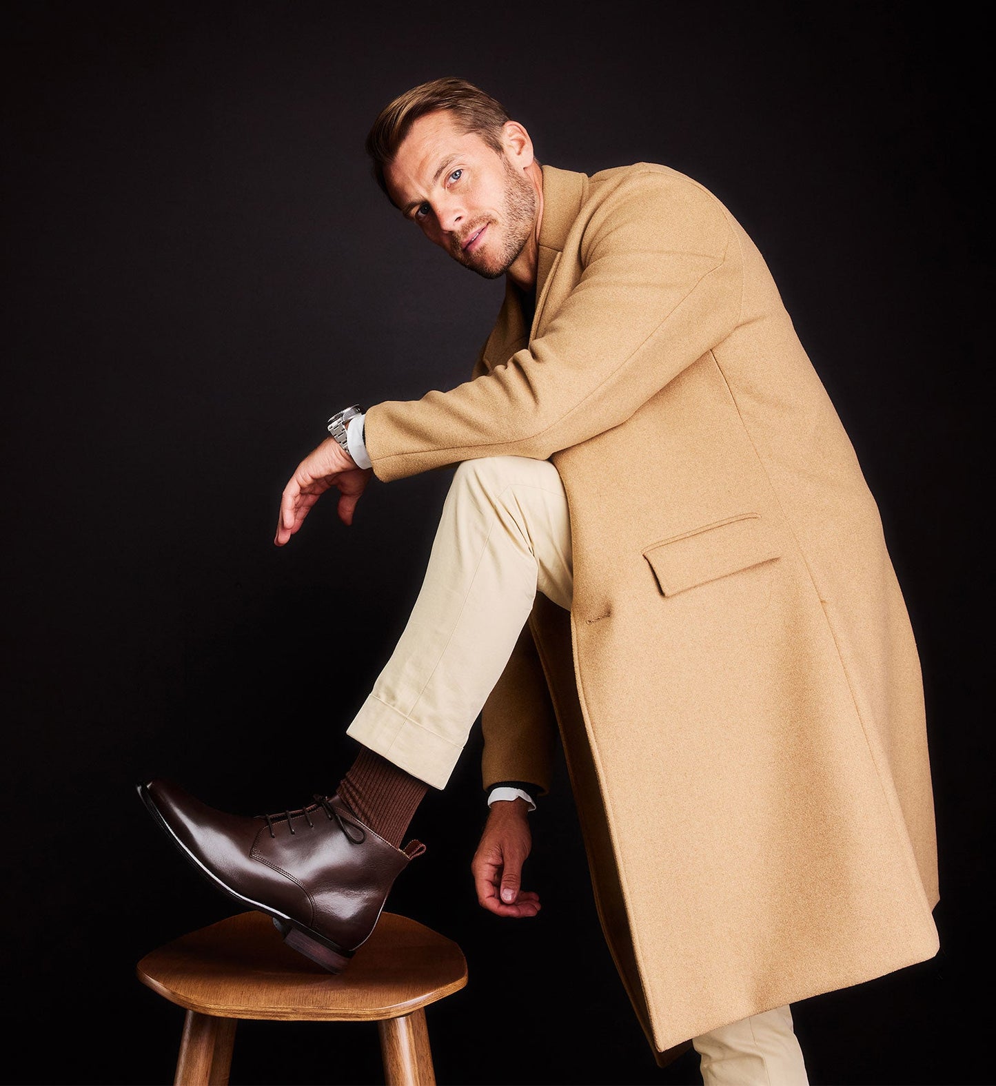 man leaning on a stool wearing low brown boots and a tan coat