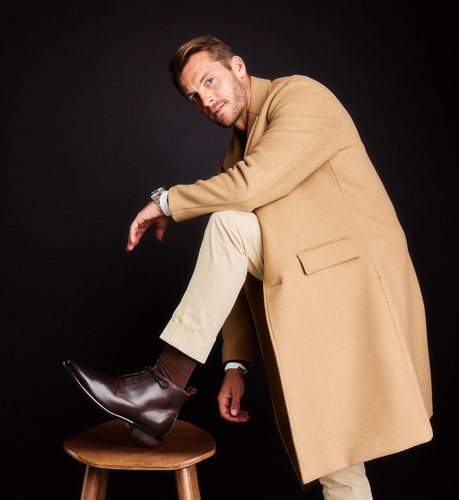 man leaning on a stool wearing low brown boots and a tan coat