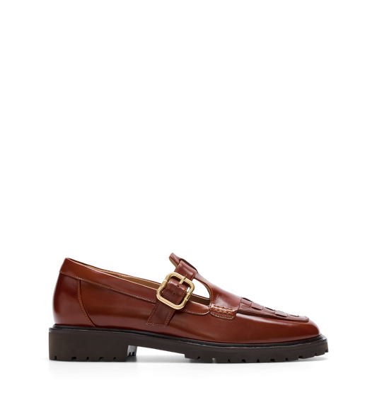 A brown leather loafer with a T-bar strap and adjustable buckle, featuring intricate brogue detailing and a chunky rubber outsole.