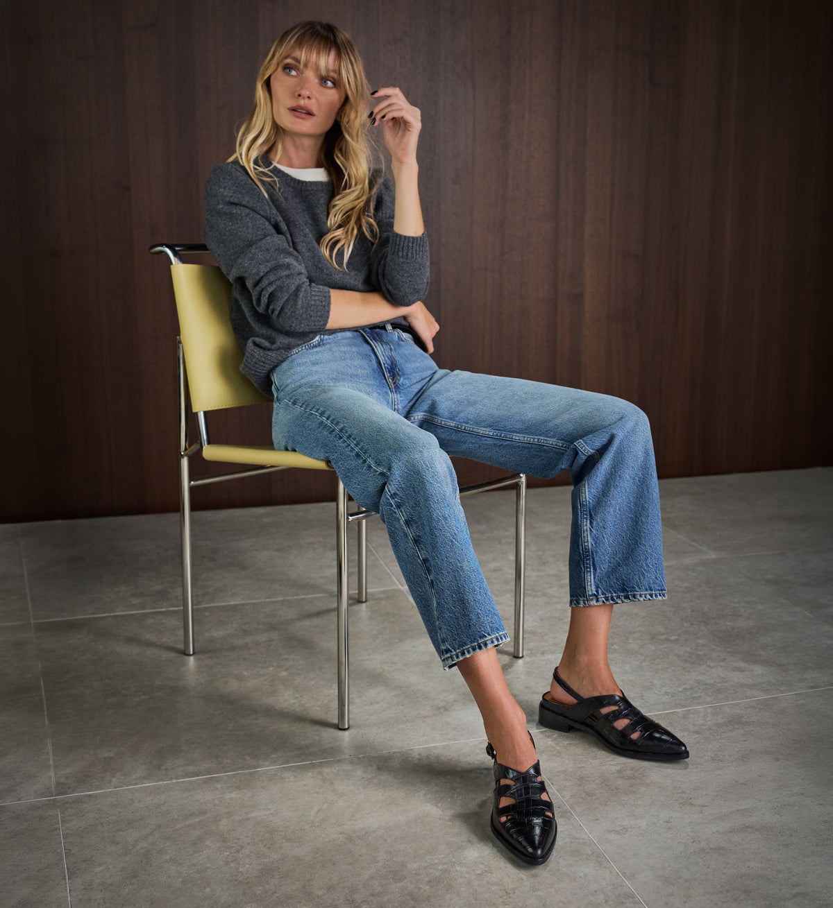 Woman sitting on a chair wearing a gray sweater, blue jeans, and black flats with cutouts