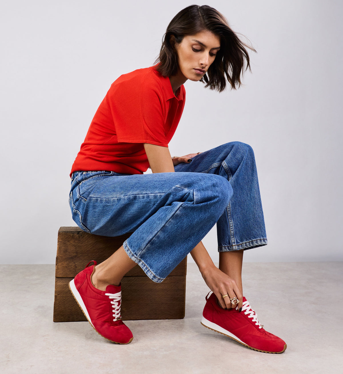 Woman wearing a red shirt, blue jeans, and red sneakers sitting on a wooden box against a plain background.