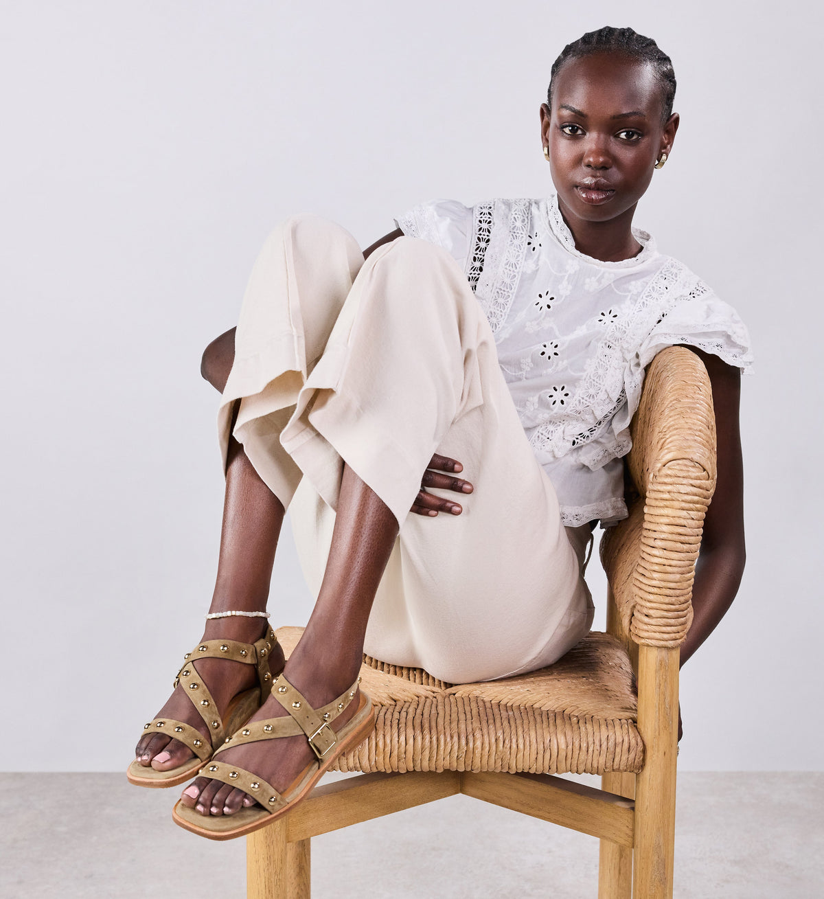 Woman sitting on a chair wearing gold studded tan sandals, white pants, and a white top with a subtle pattern.