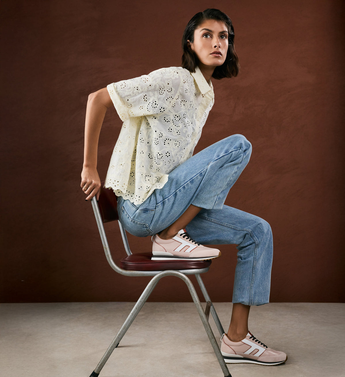 Woman sitting on a chair wearing a white blouse, blue jeans, and pink sneakers against a brown background