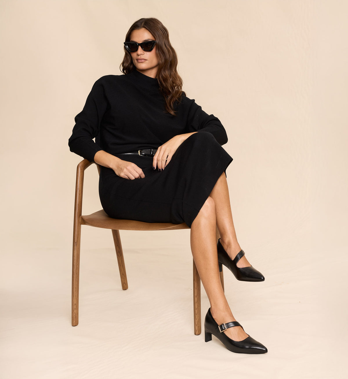 Woman in a black outfit with black pointed toe heels sitting on a wooden chair against a beige background