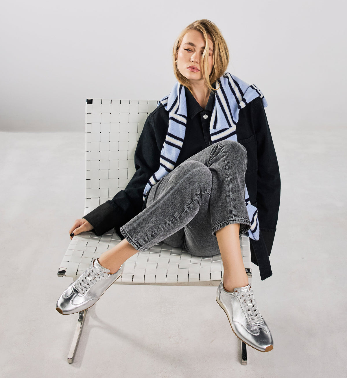 Woman sitting on a chair wearing a black jacket, gray jeans, and silver sneakers with a striped scarf.
