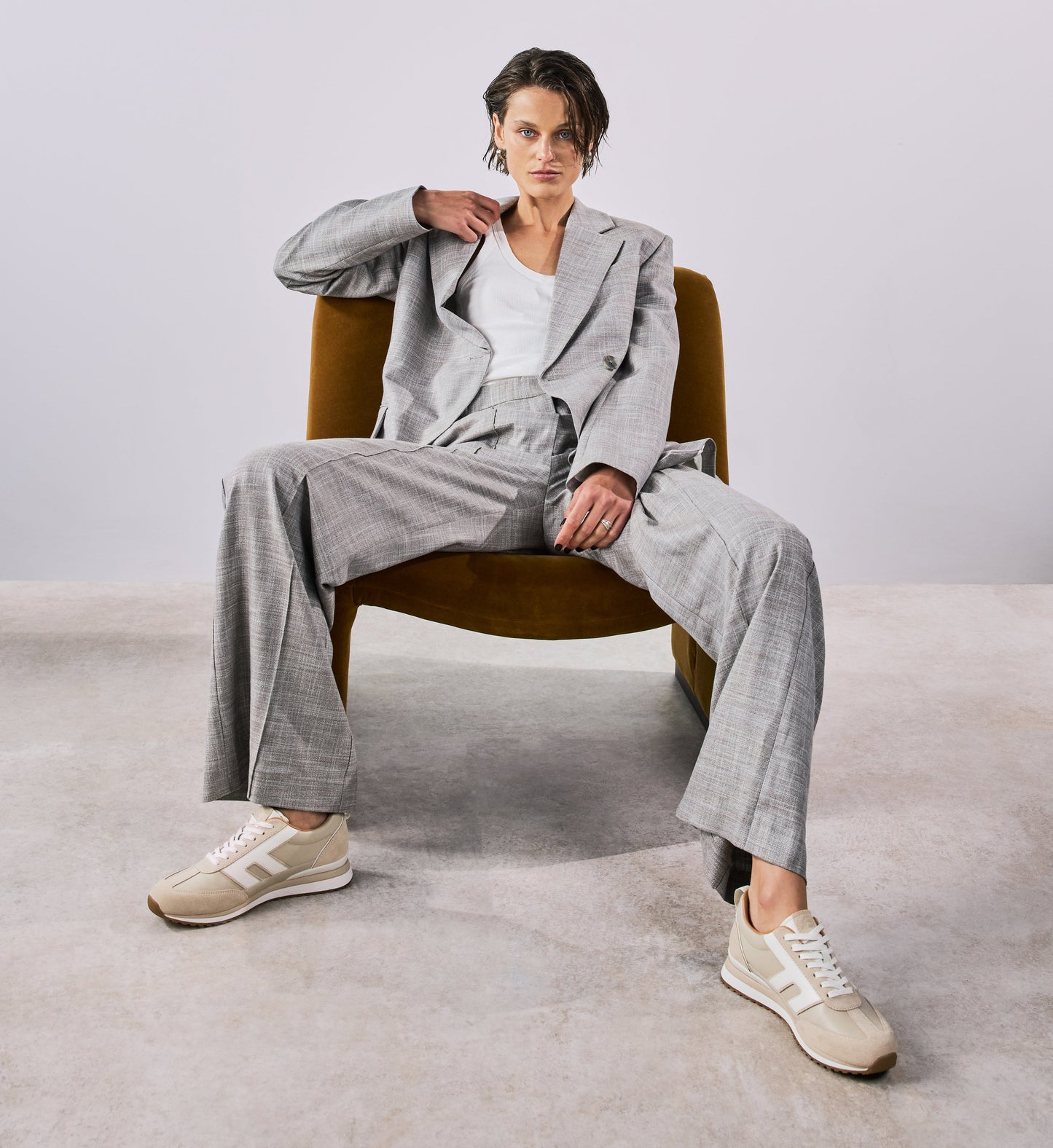 Person wearing a gray suit sitting on a chair with beige sneakers a plain background