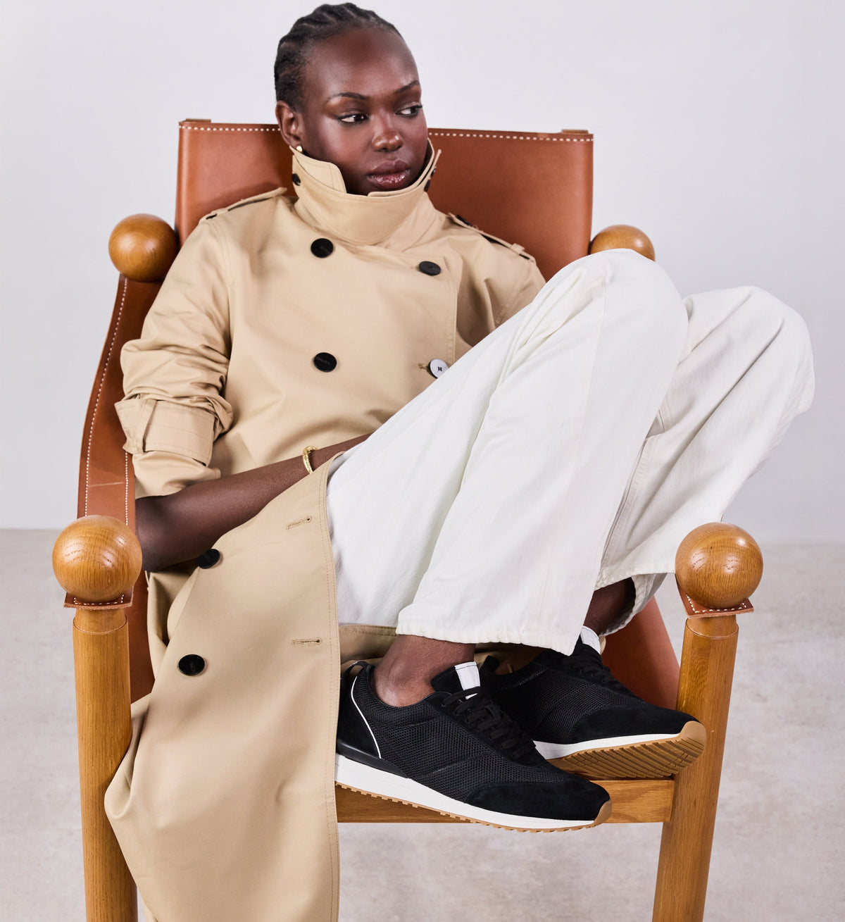 Person wearing a beige trench coat and white pants sitting on a wooden chair.