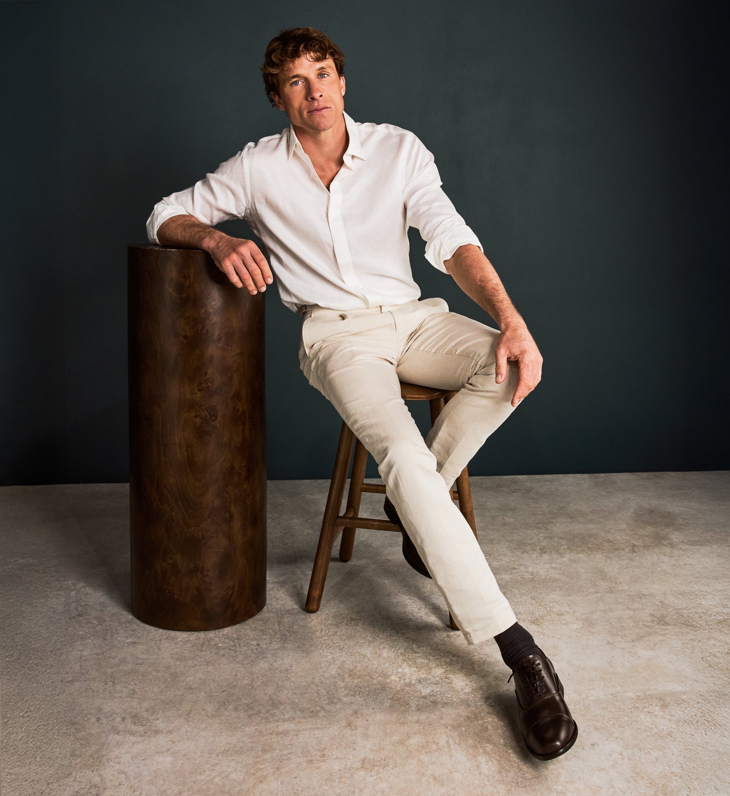 Man sitting on a wooden stool next to a dark cylindrical object against a gray background