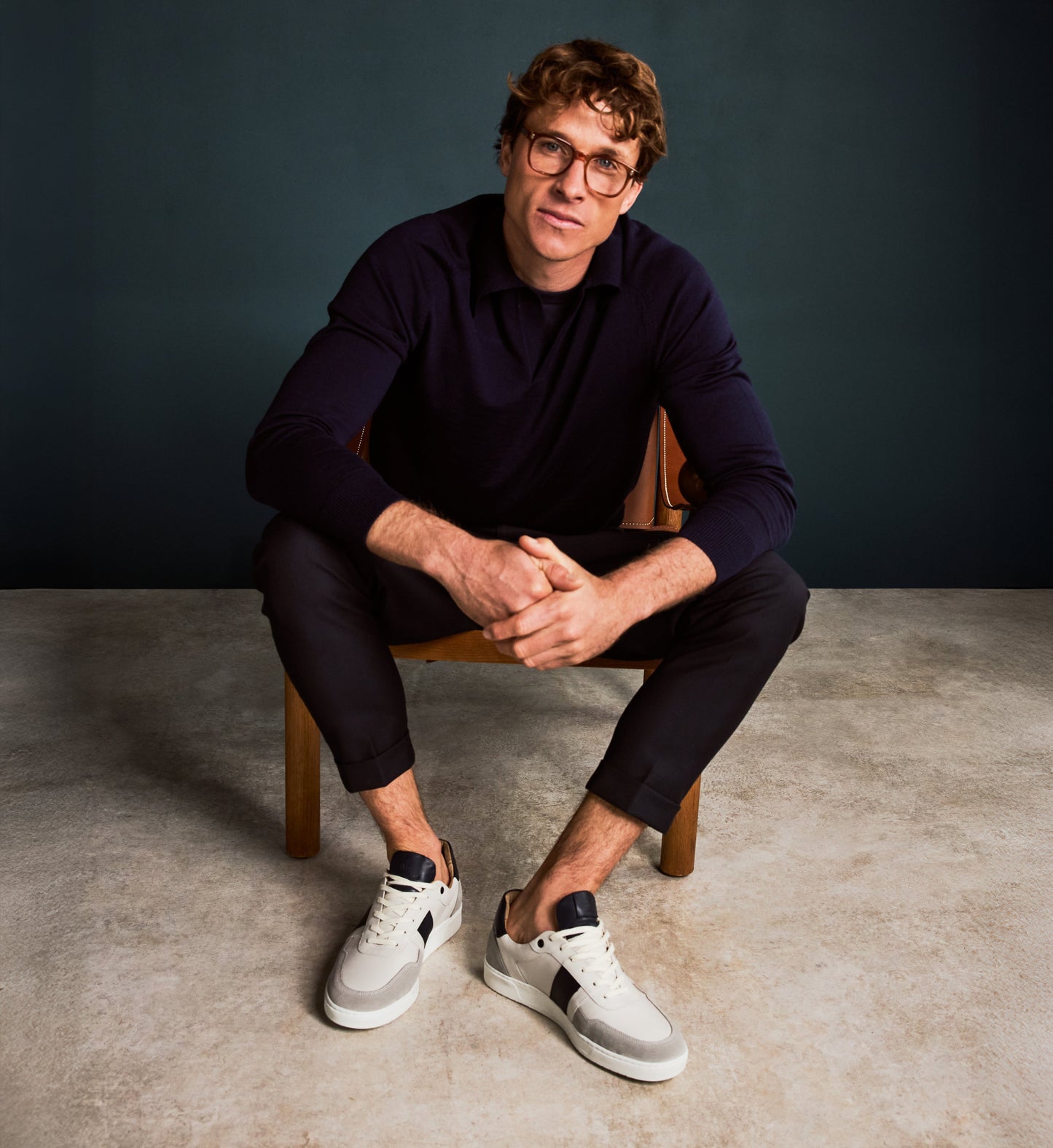 Man sitting on a chair wearing glasses, a dark sweater, black pants, and white sneakers with navy stripes