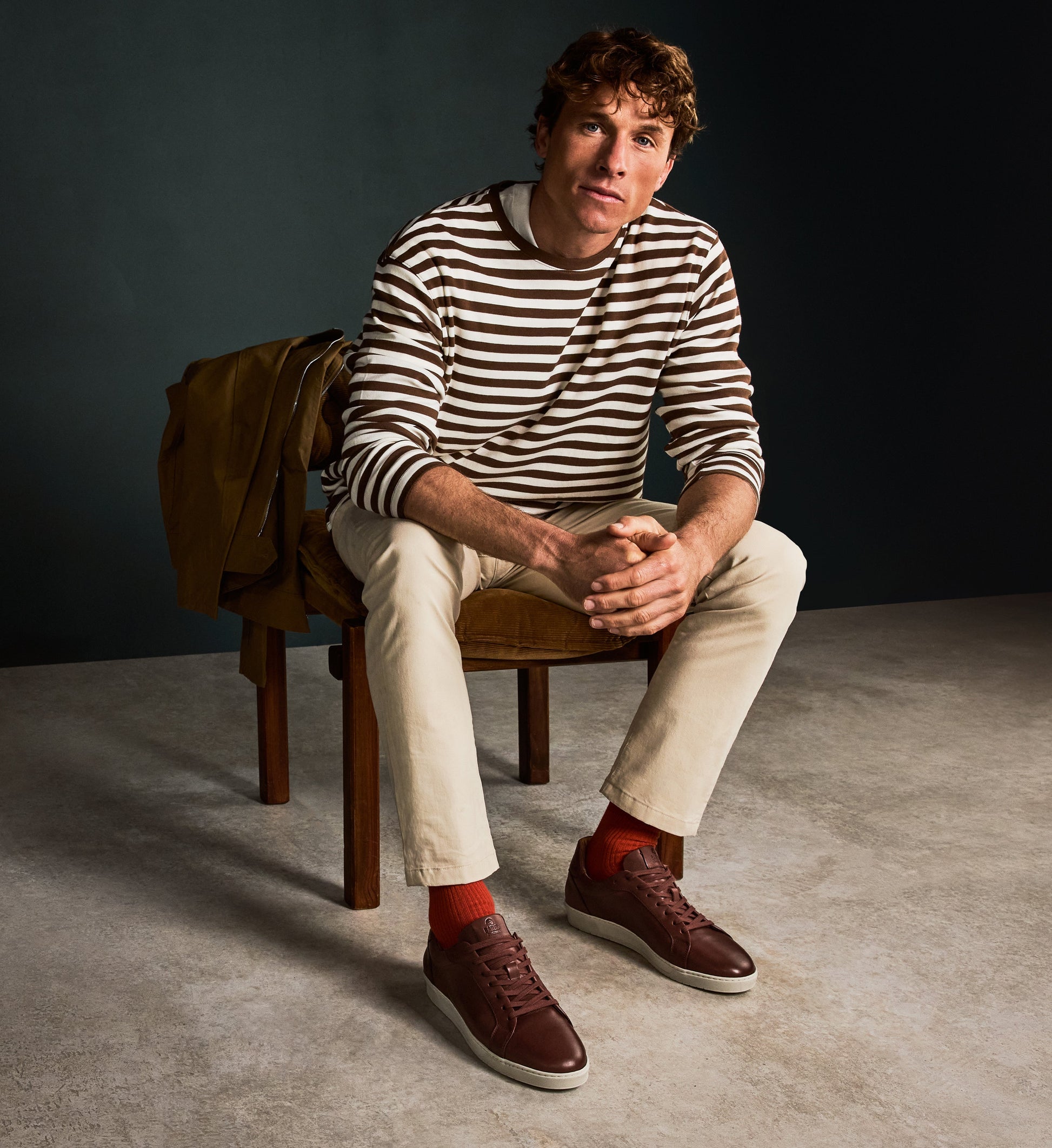 Man sitting on a chair wearing a striped sweater and beige pants with a dark background and brown sneakers