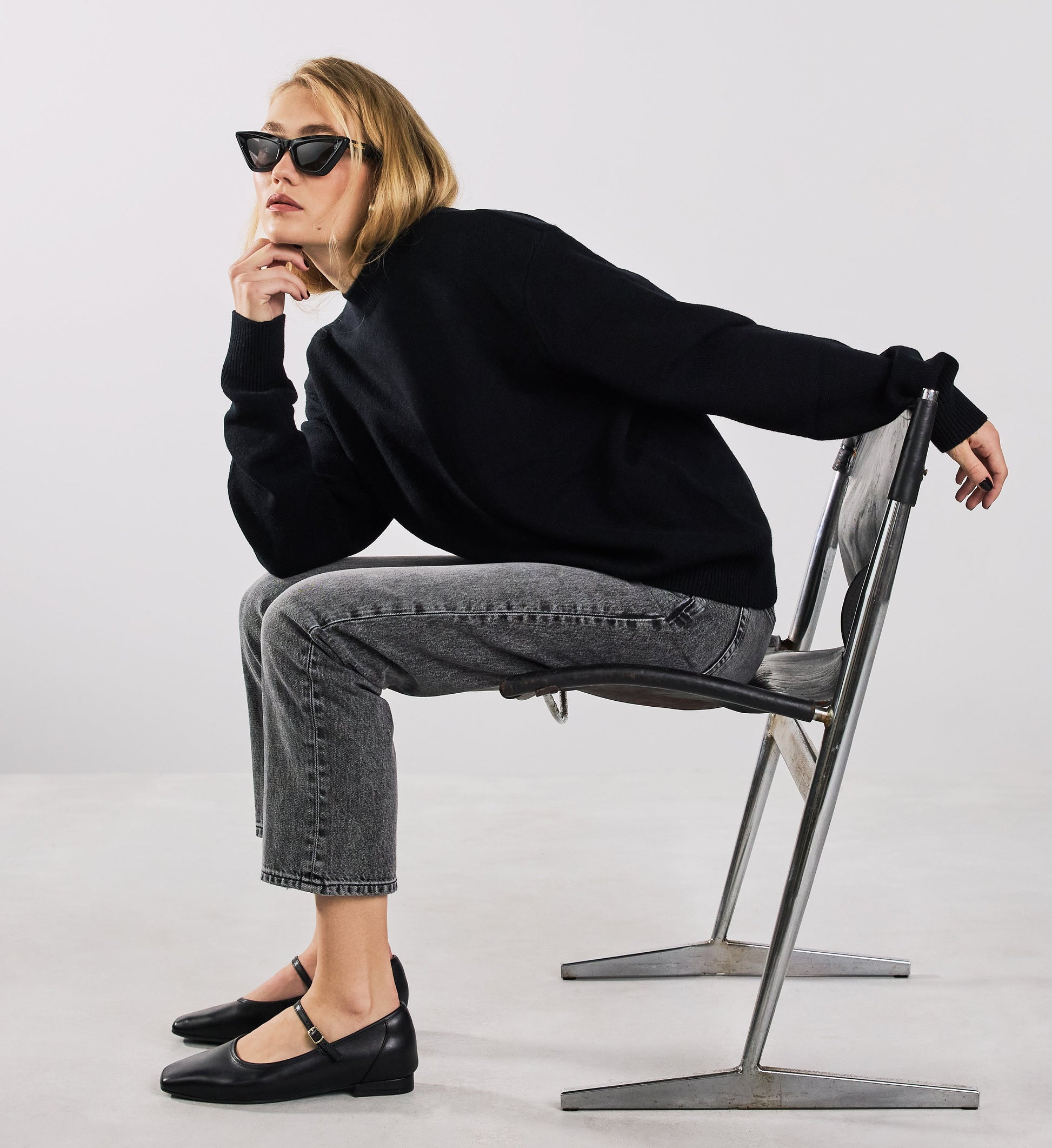 Person wearing a black sweater and gray jeans, plus bared footwear ballet flats sitting on a chair against a white background