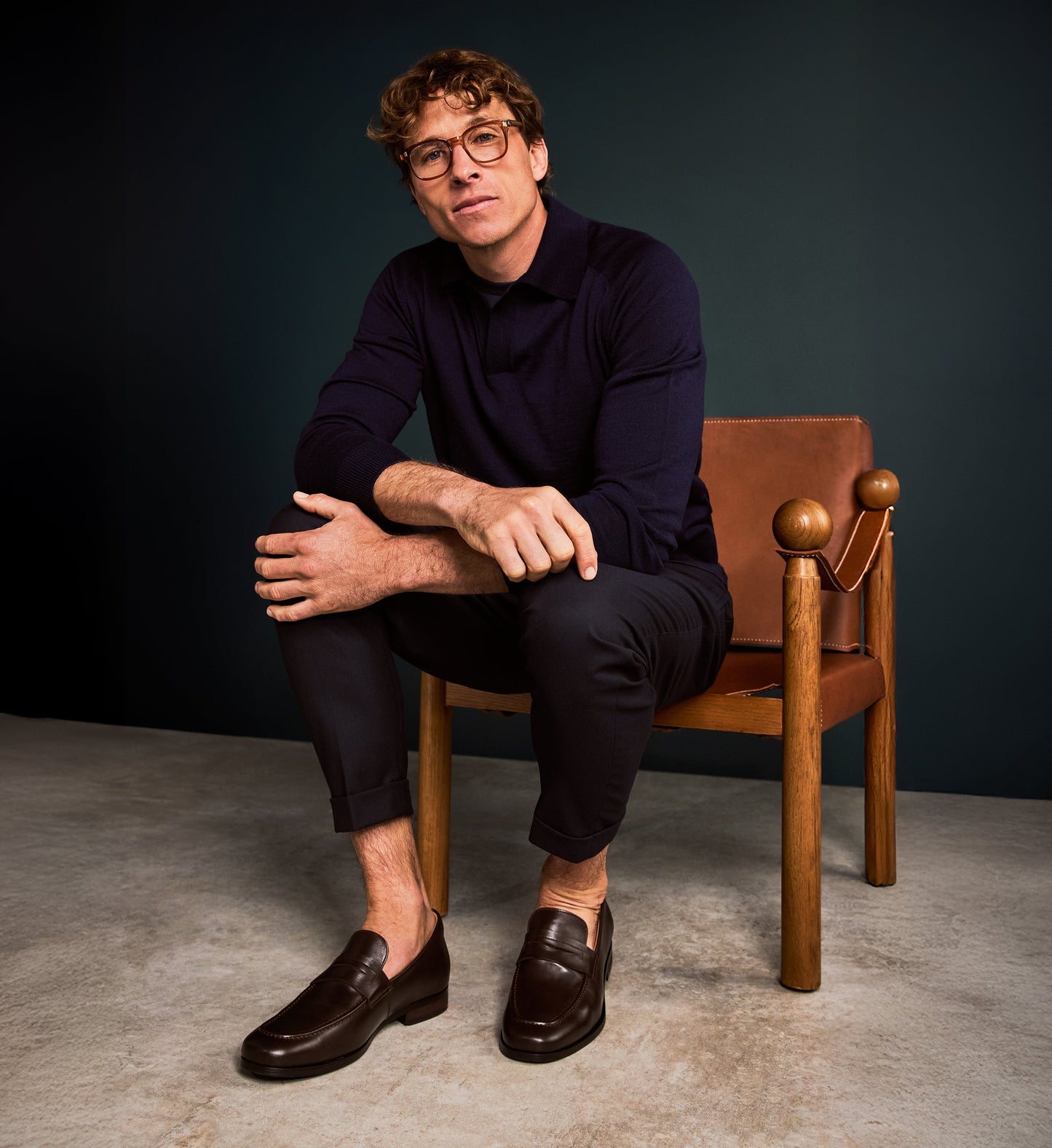 Man sitting on a brown chair against a dark background wearing brown loafers and a navy polo shirt