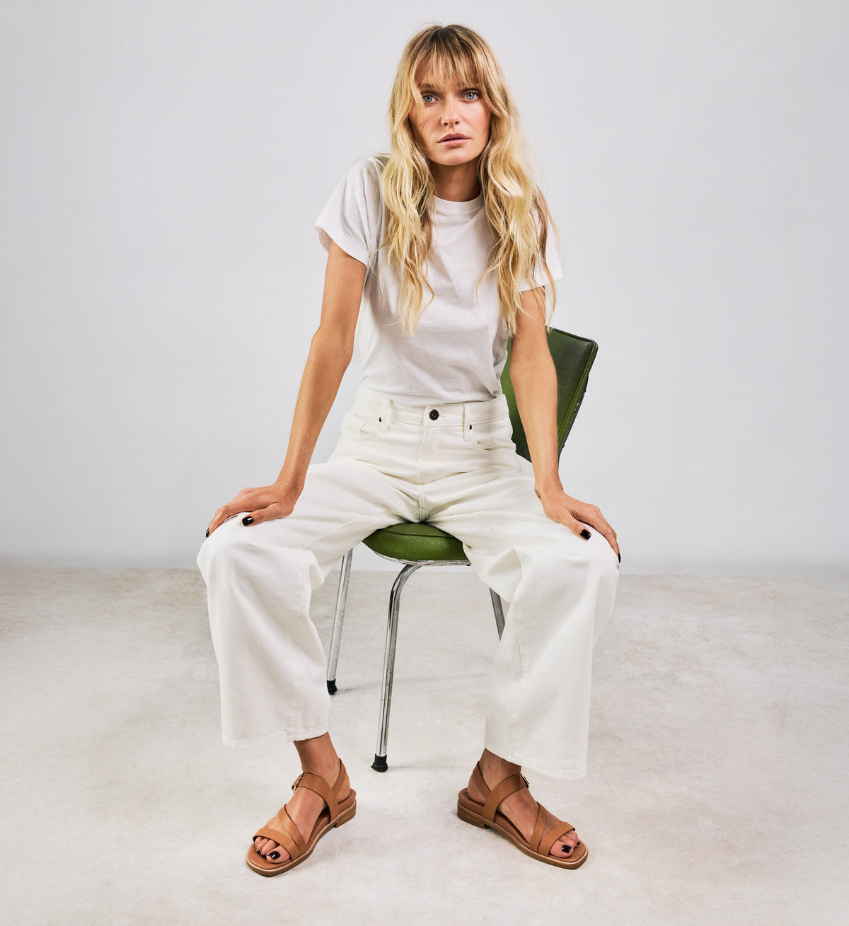 Woman sitting on a green chair wearing a white t-shirt and white pants against a plain background with strappy tan sandals