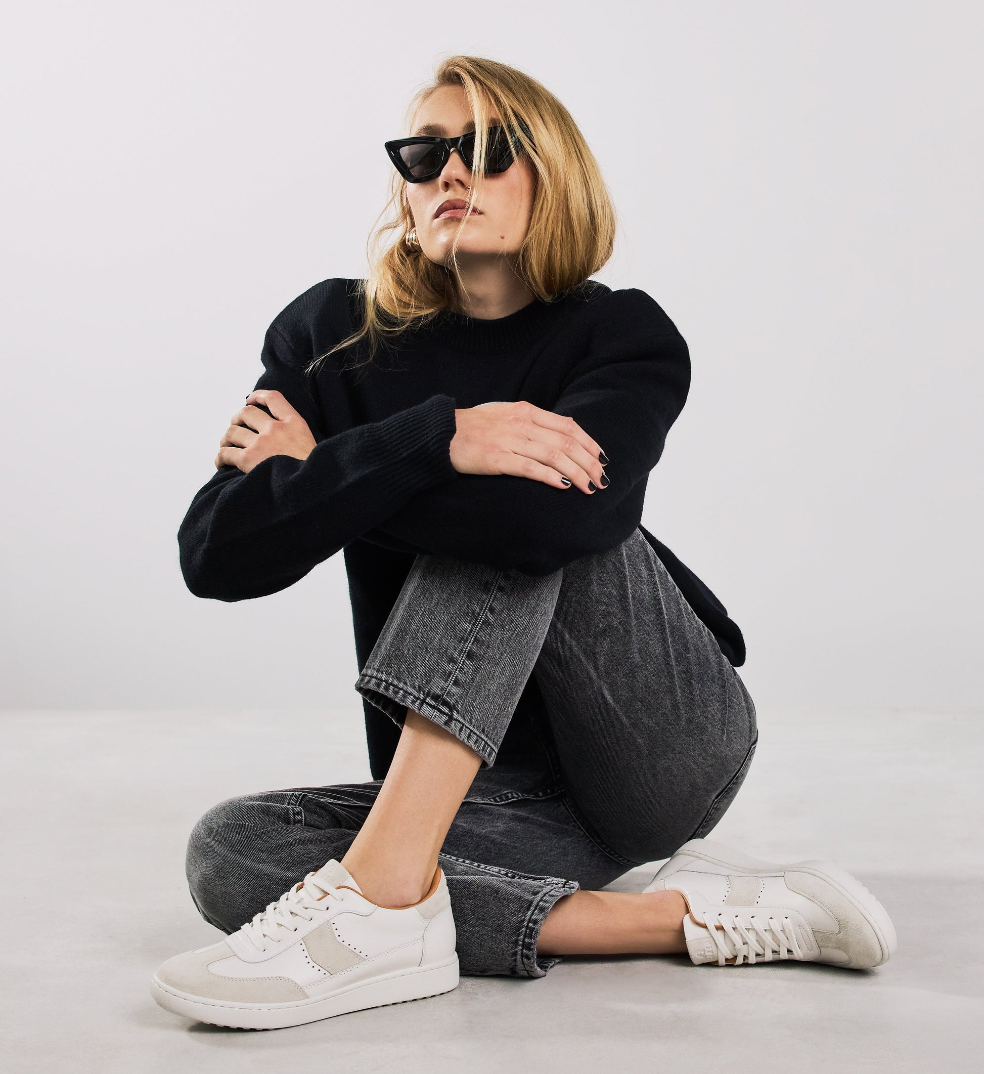Woman wearing a black sweater, gray jeans, and white sneakers sitting on a light-colored floor.
