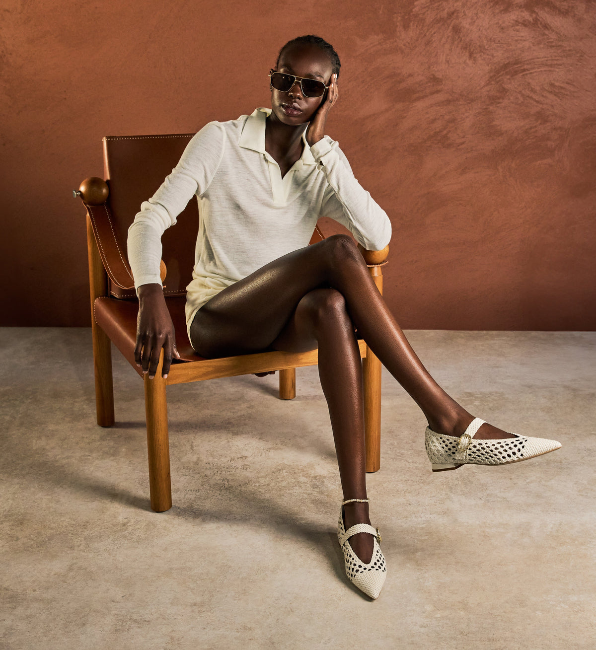 Woman sitting on a wooden chair wearing sunglasses, a white shirt, black tights, and patterned shoes against a brown background.