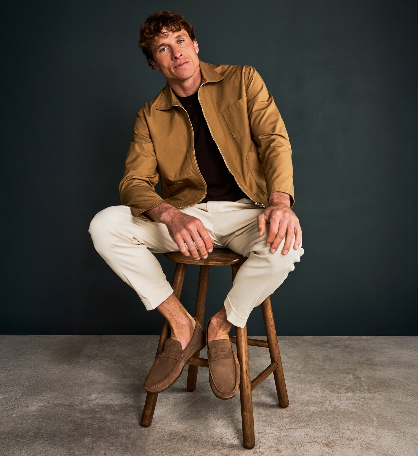 Man sitting on a wooden stool wearing a brown jacket, white pants, and brown suede shoes against a dark background.
