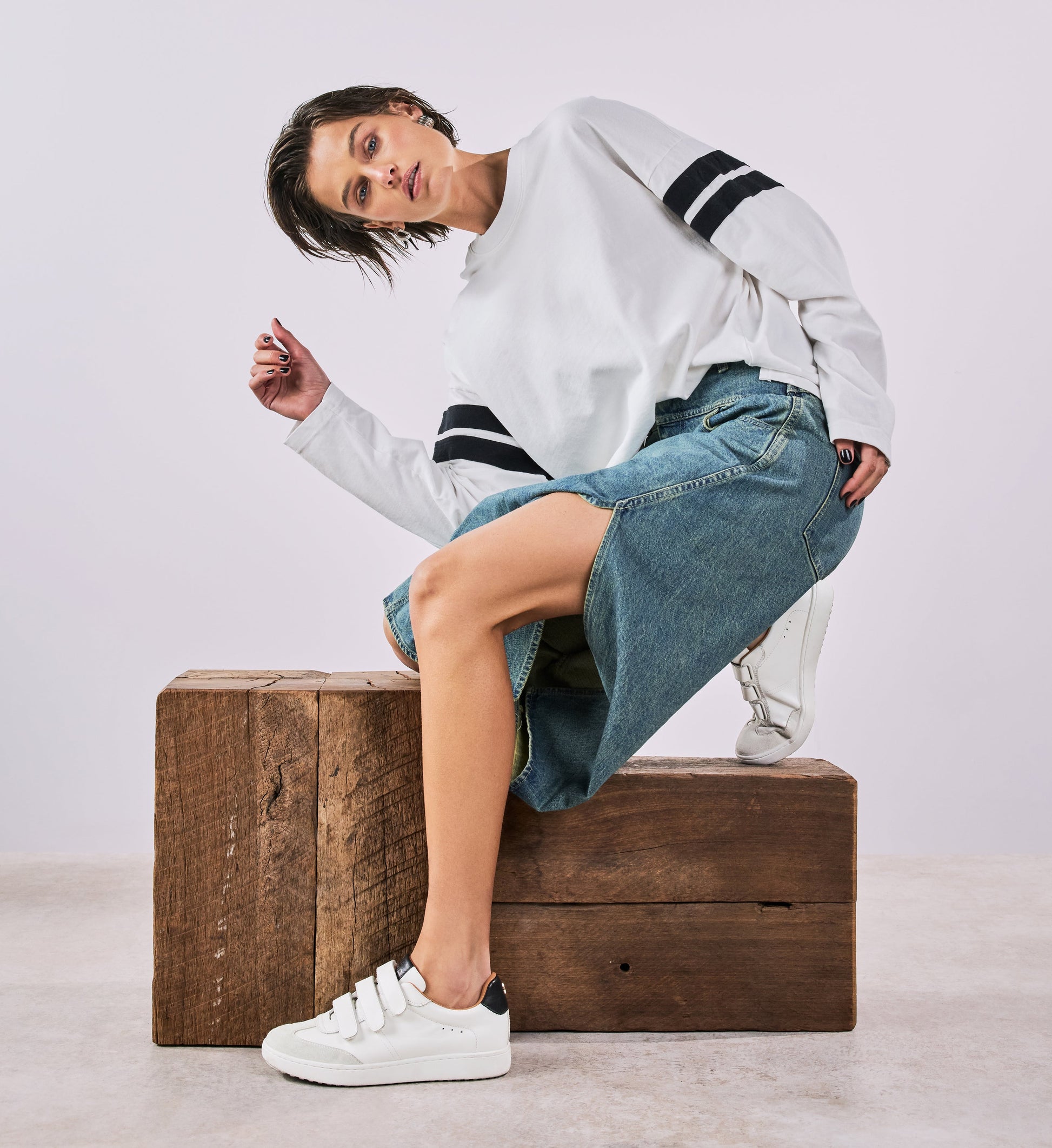Person sitting on a wooden block wearing a white sweatshirt, blue jeans, and white sneakers.