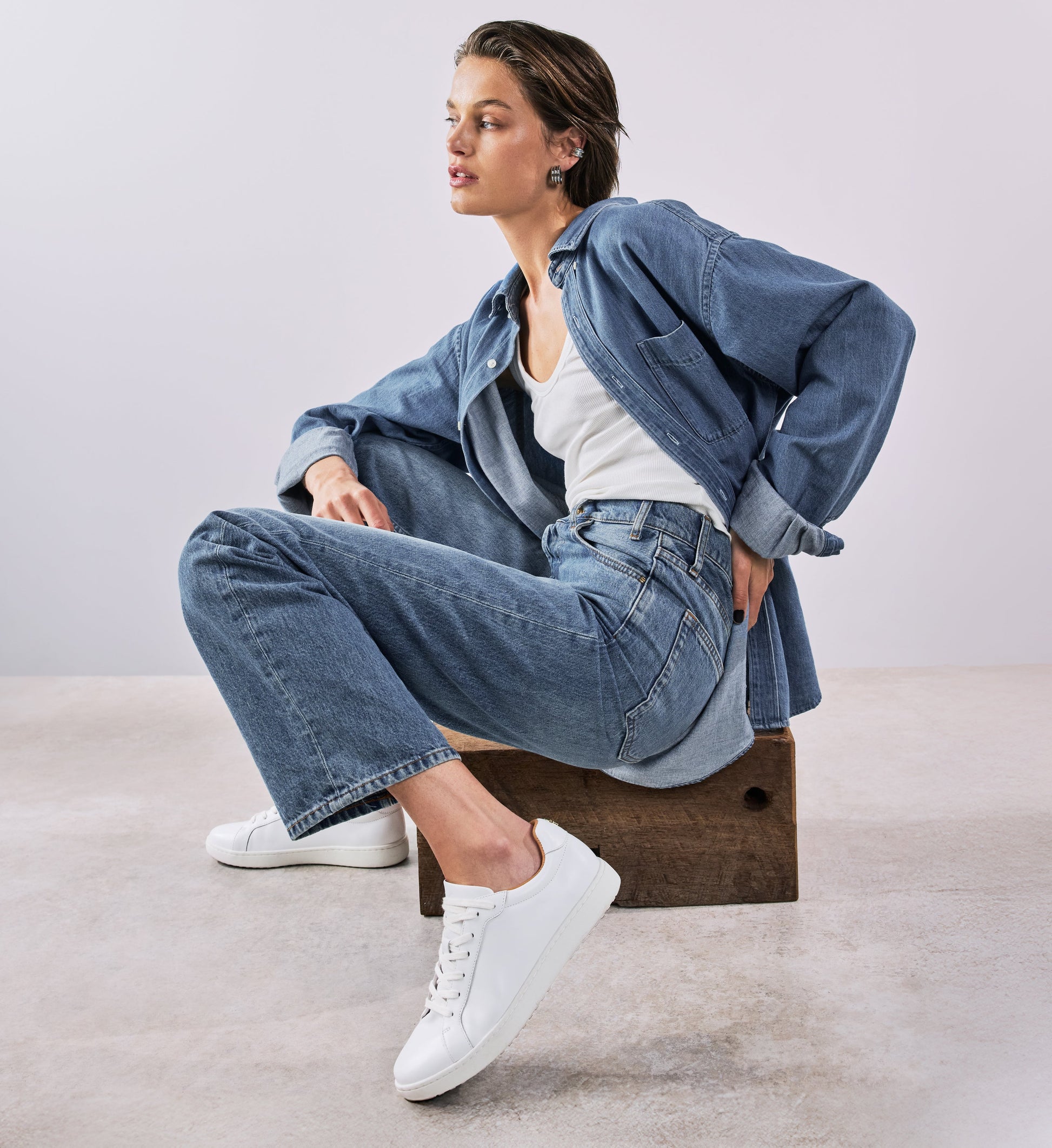 Woman wearing a denim outfit with white sneakers sitting on a wooden block.